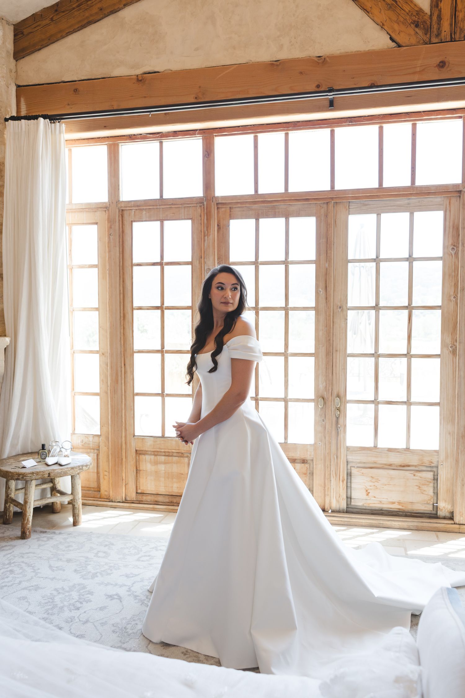 A bride in a classic white wedding gown poses by large windows in a rustic room with wooden beams.