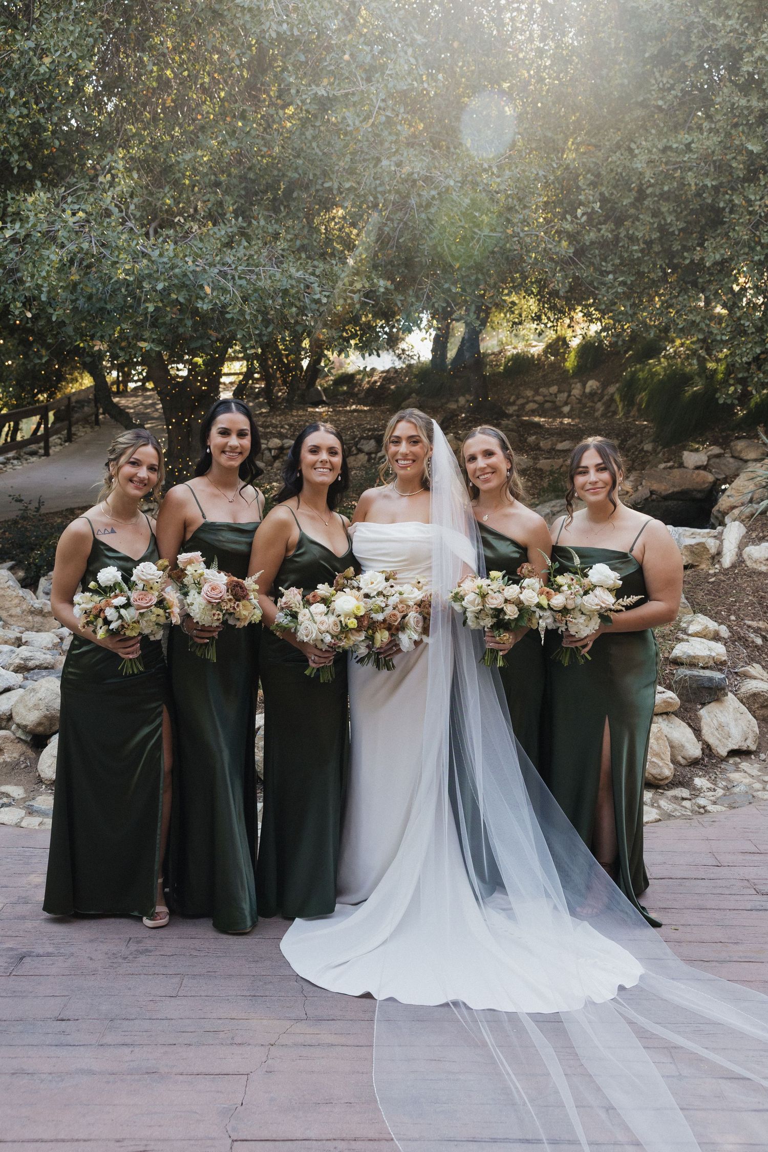 Bridesmaids in elegant dark green dresses pose with the bride holding white floral bouquets outdoors.