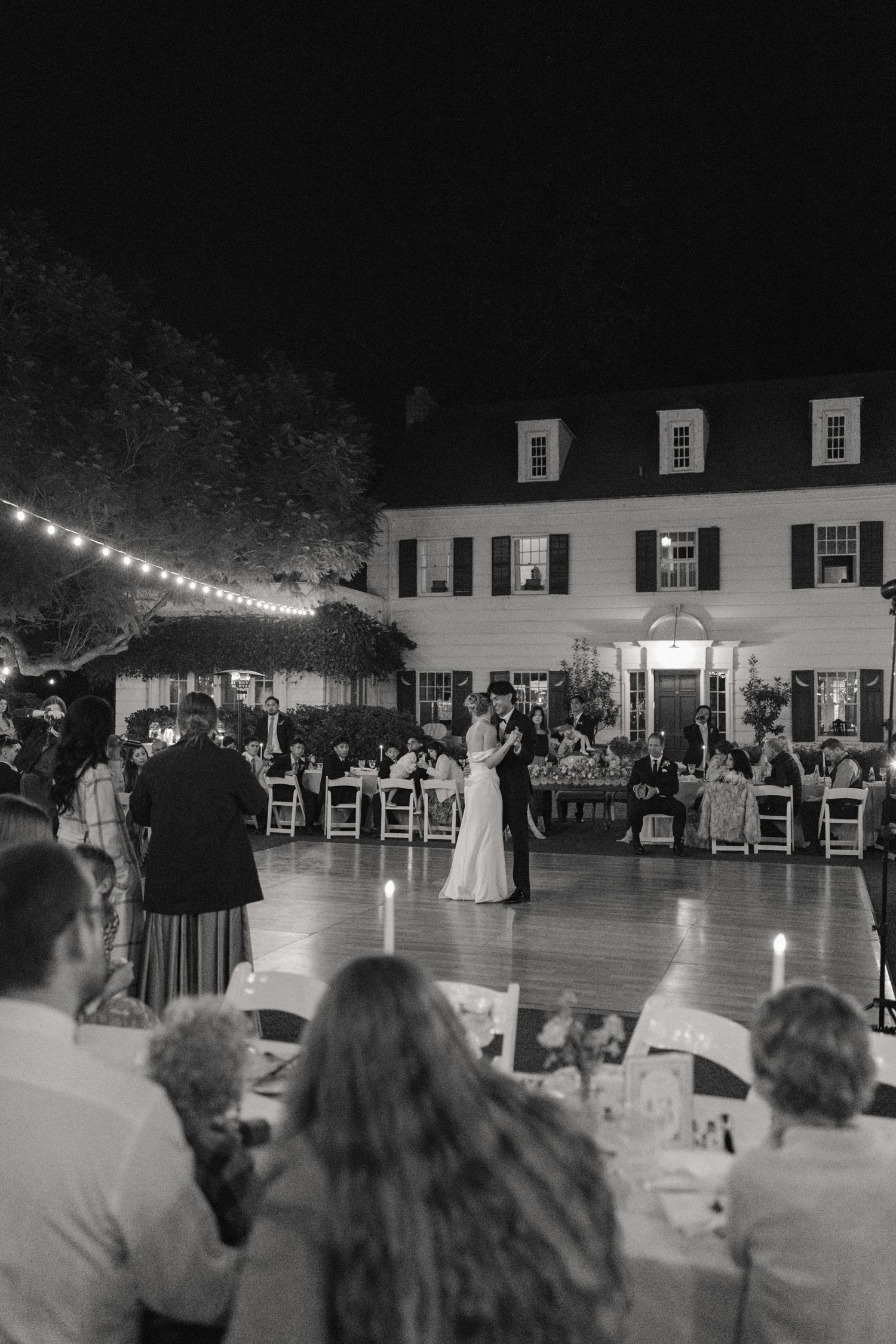 A black and white wedding reception photo showing couples dancing on a patio with string lights at a historic house venue.