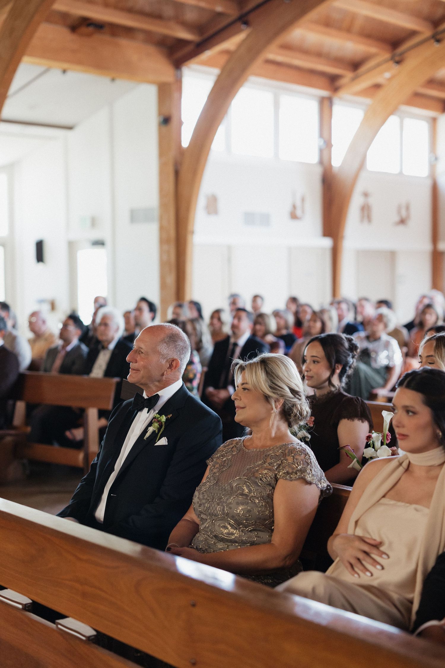 Wedding guests seated in wooden church pews during a ceremony in a bright venue with timber beam architecture.
