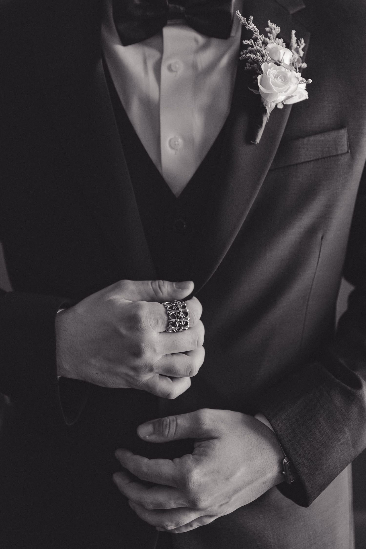 Black and white photo of groom in formal suit wearing boutonniere and ornate ring.