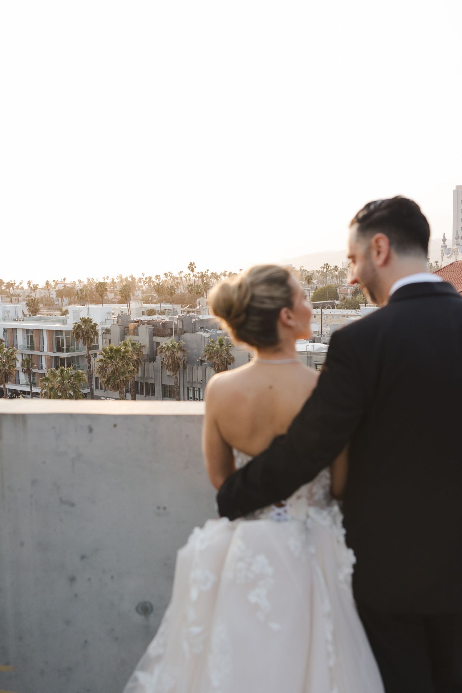 Bride and groom share an intimate moment overlooking a cityscape at sunset from a rooftop venue.