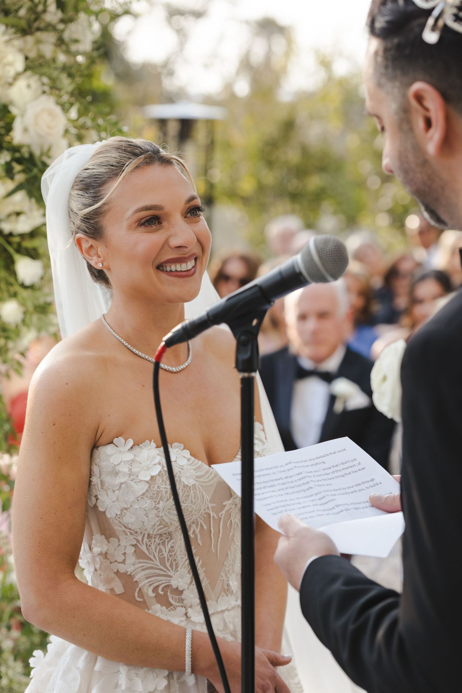 Couple exchanging vows at outdoor wedding ceremony with microphone and floral arch in background during sunny day.