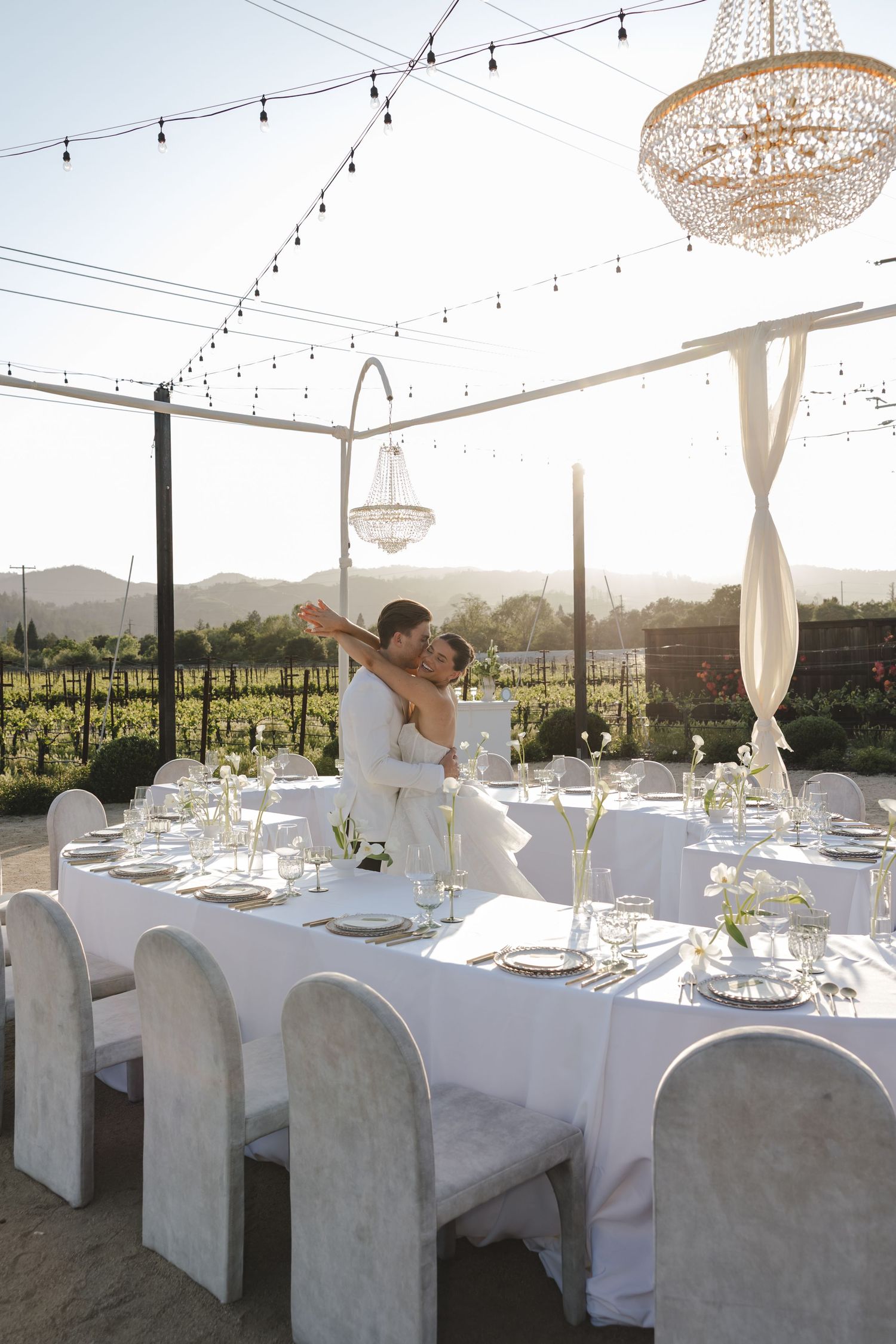 Elegant outdoor wedding reception with white table settings, crystal chandeliers and string lights overlooking vineyard at sunset.