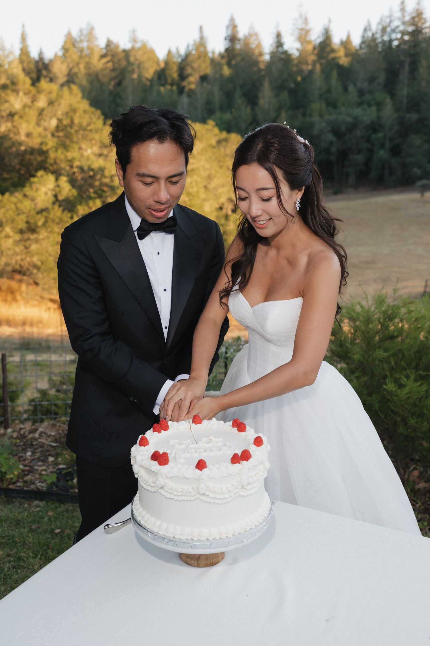A newlywed couple cuts into their white wedding cake decorated with red hearts outdoors.