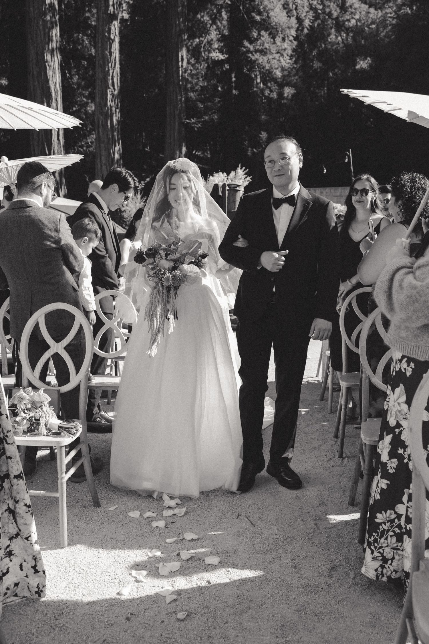 A wedding ceremony in a forest setting with guests seated on white chairs as a couple walks down the aisle.