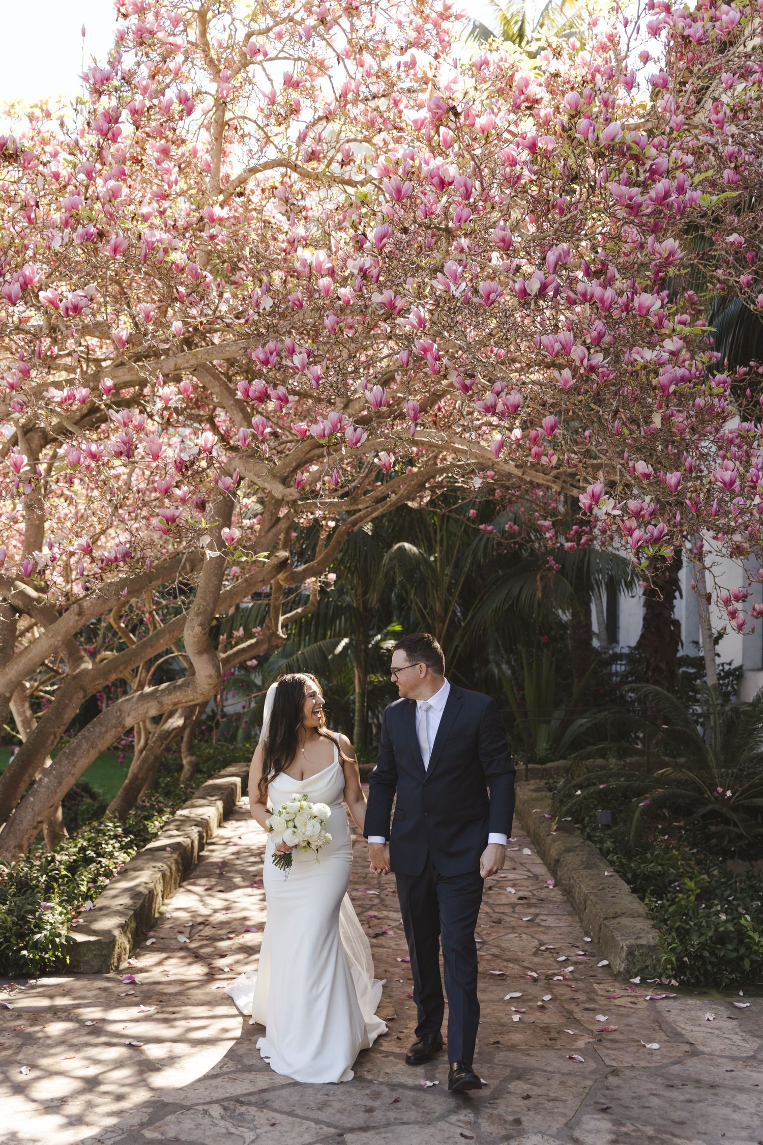 Couple walking together under blooming pink magnolia tree in garden pathway during spring wedding.