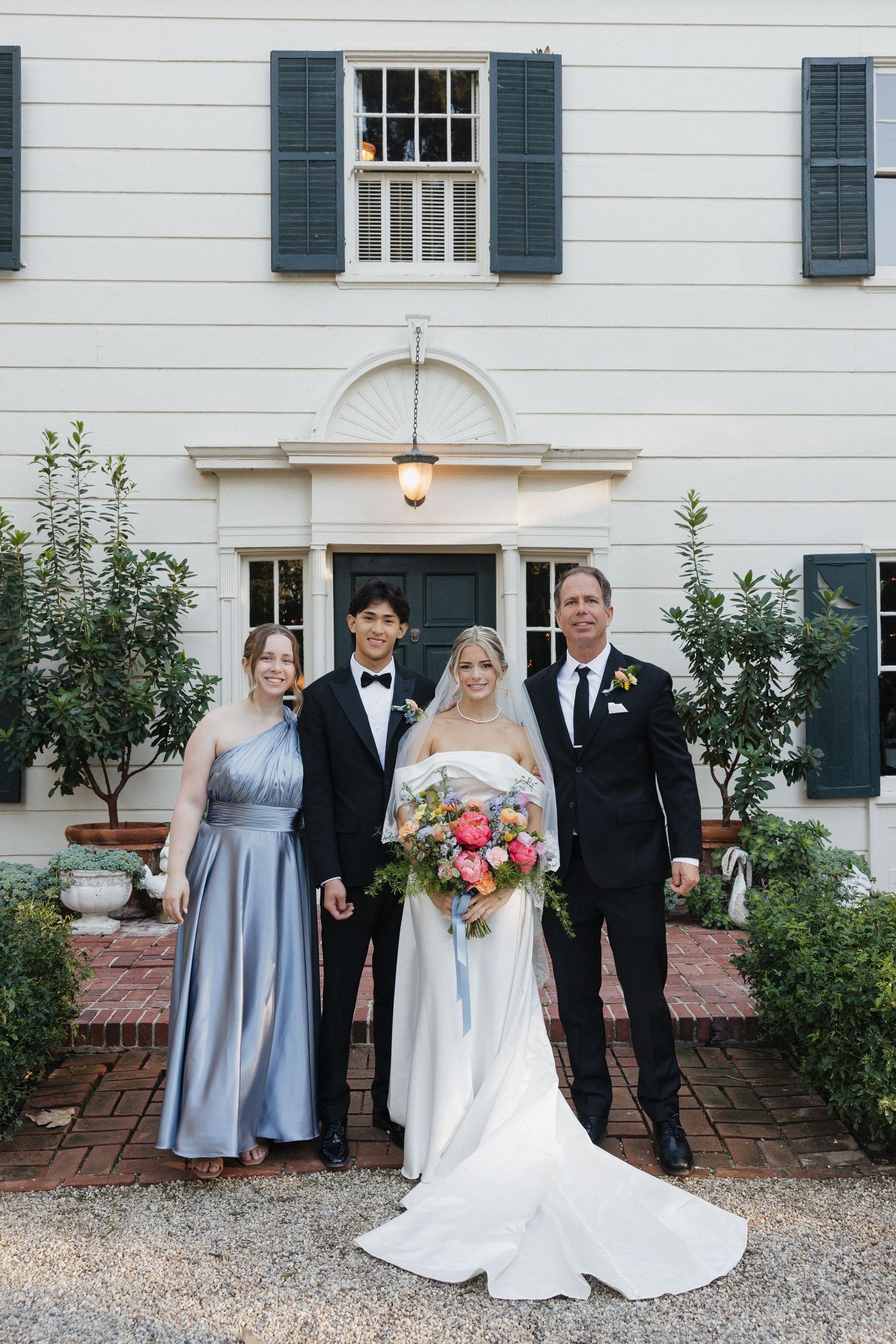 Wedding party poses in front of a classic white colonial house with black shutters and a brick walkway.