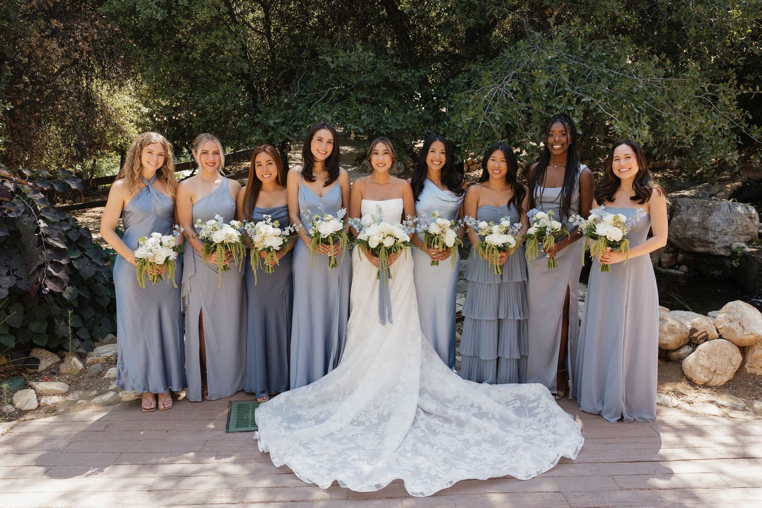 Bridal party in matching gray dresses poses outdoors with bride in lace gown.