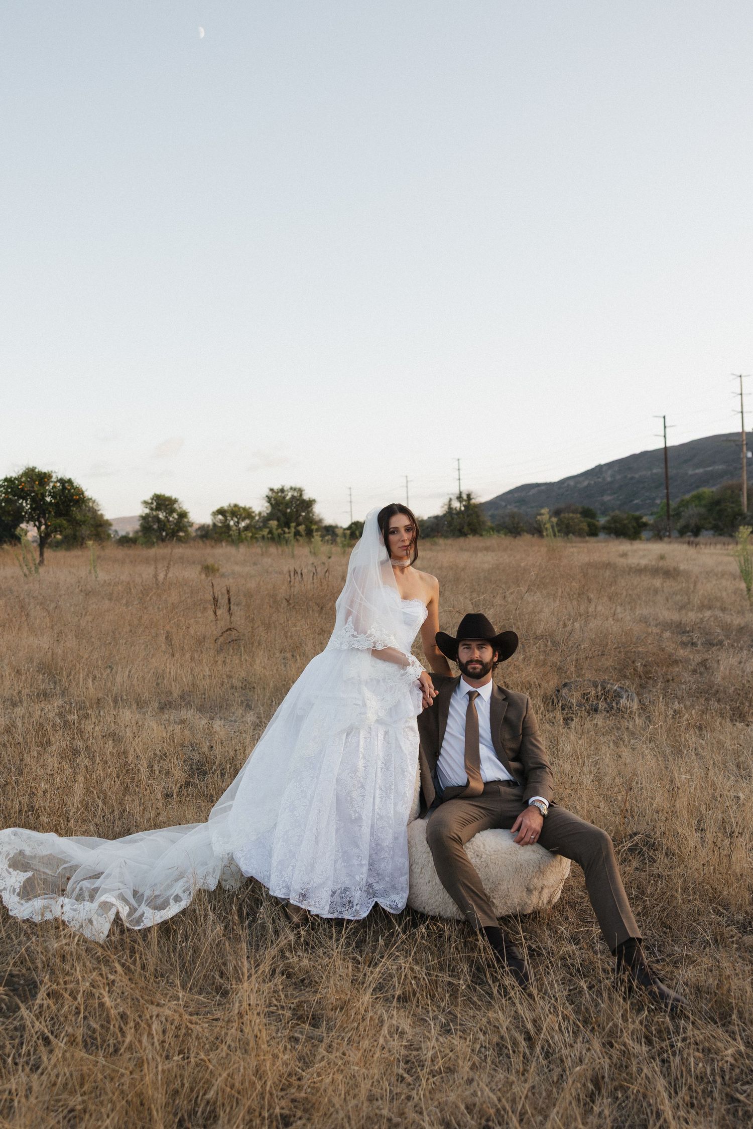 A couple in wedding attire sits together in a rustic field of golden grass at sunset.