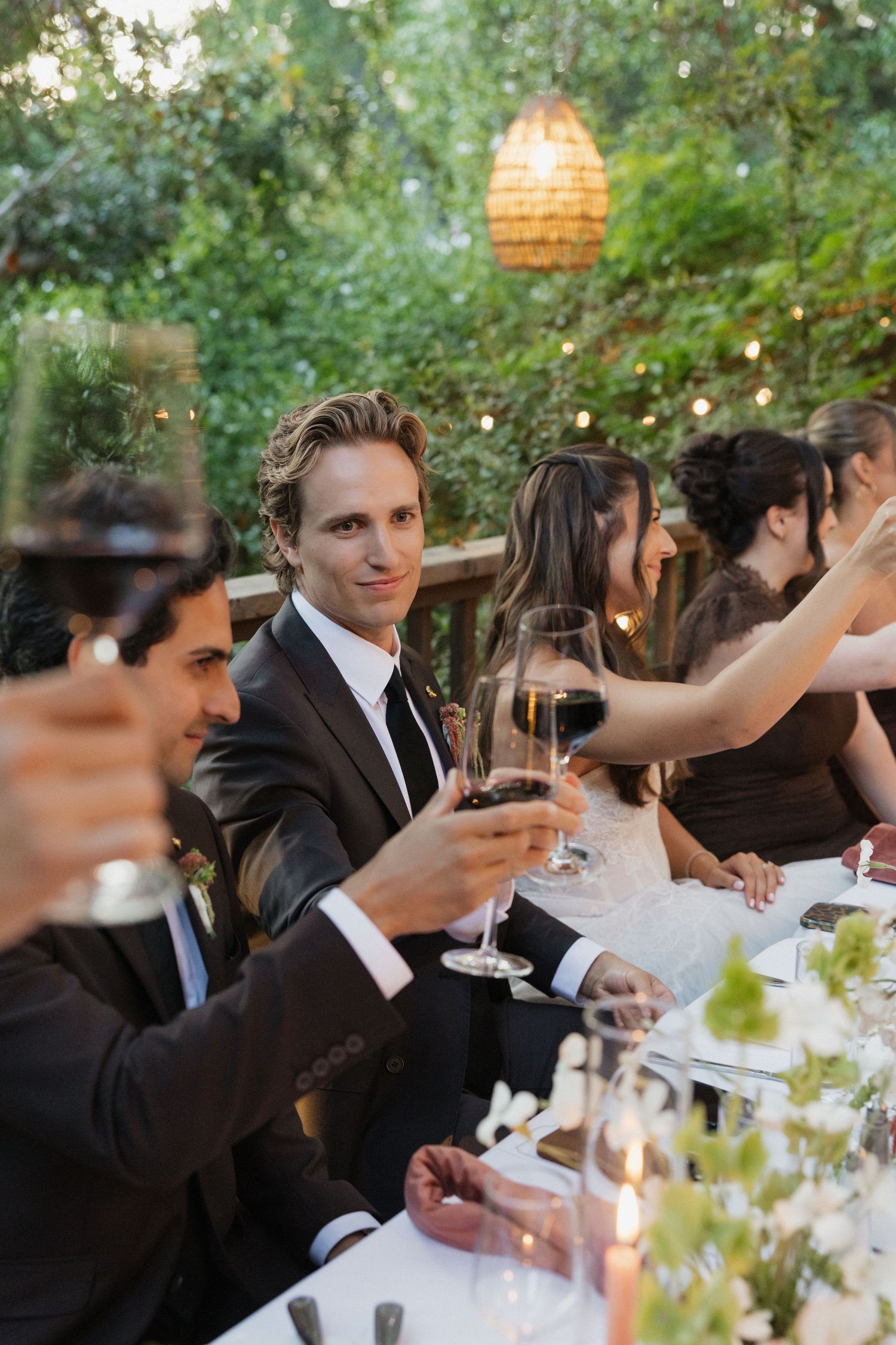 Groom in a dark suit raises a wine glass during an outdoor wedding reception dinner.