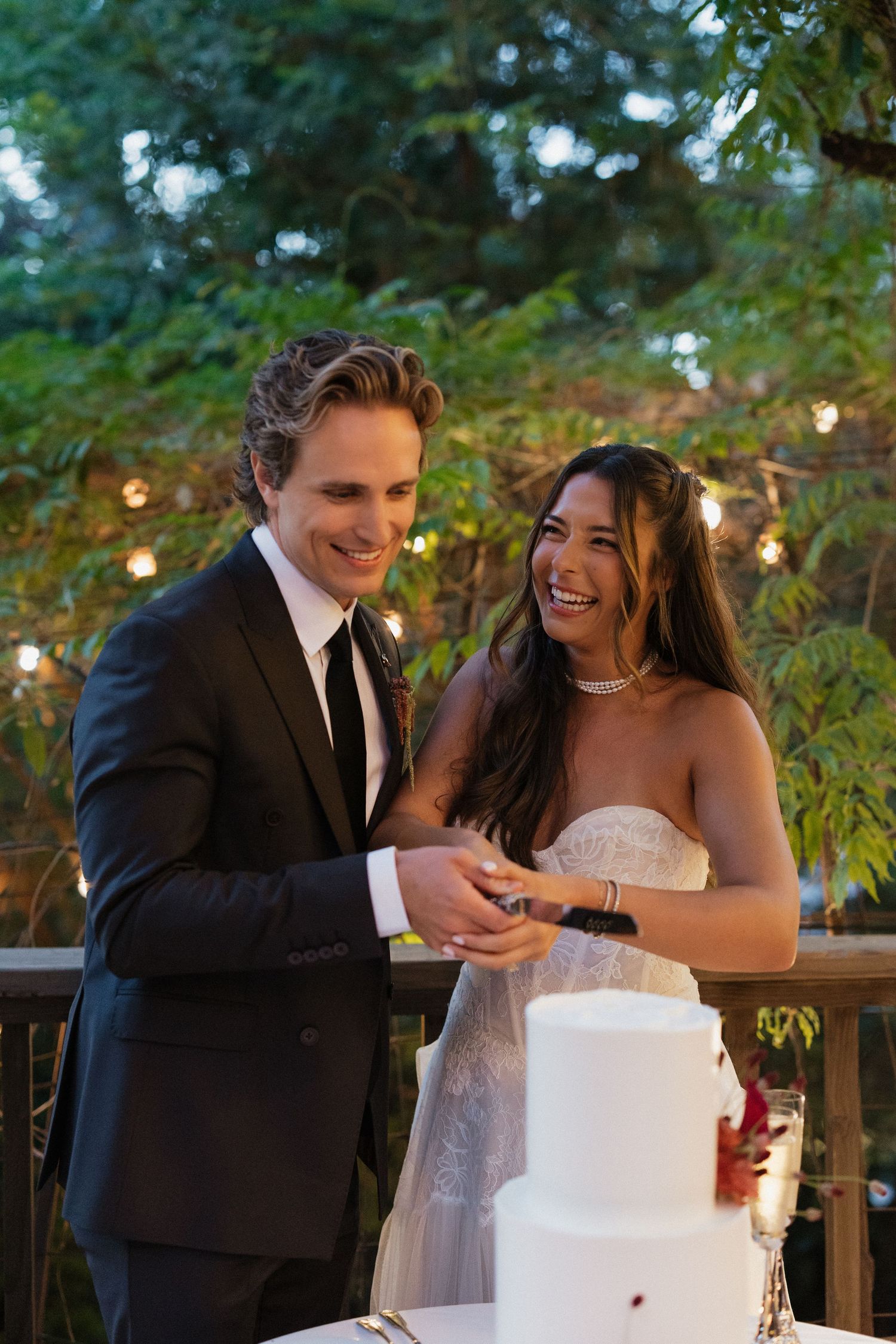 Laughing newlyweds cut elegant white wedding cake outdoors at evening reception under string lights.