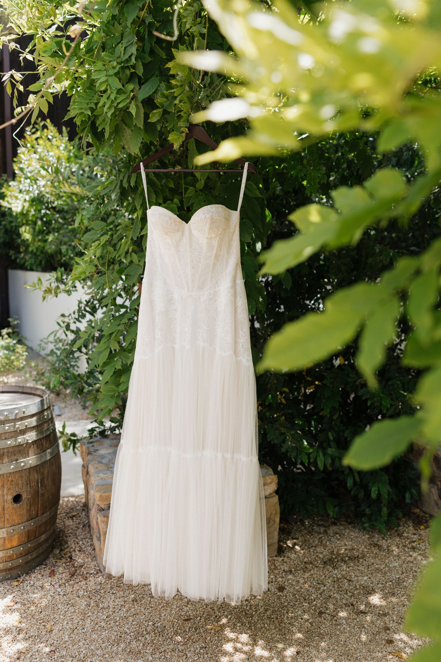 Elegant strapless white tulle wedding dress hanging outdoors among green foliage near wooden barrels.