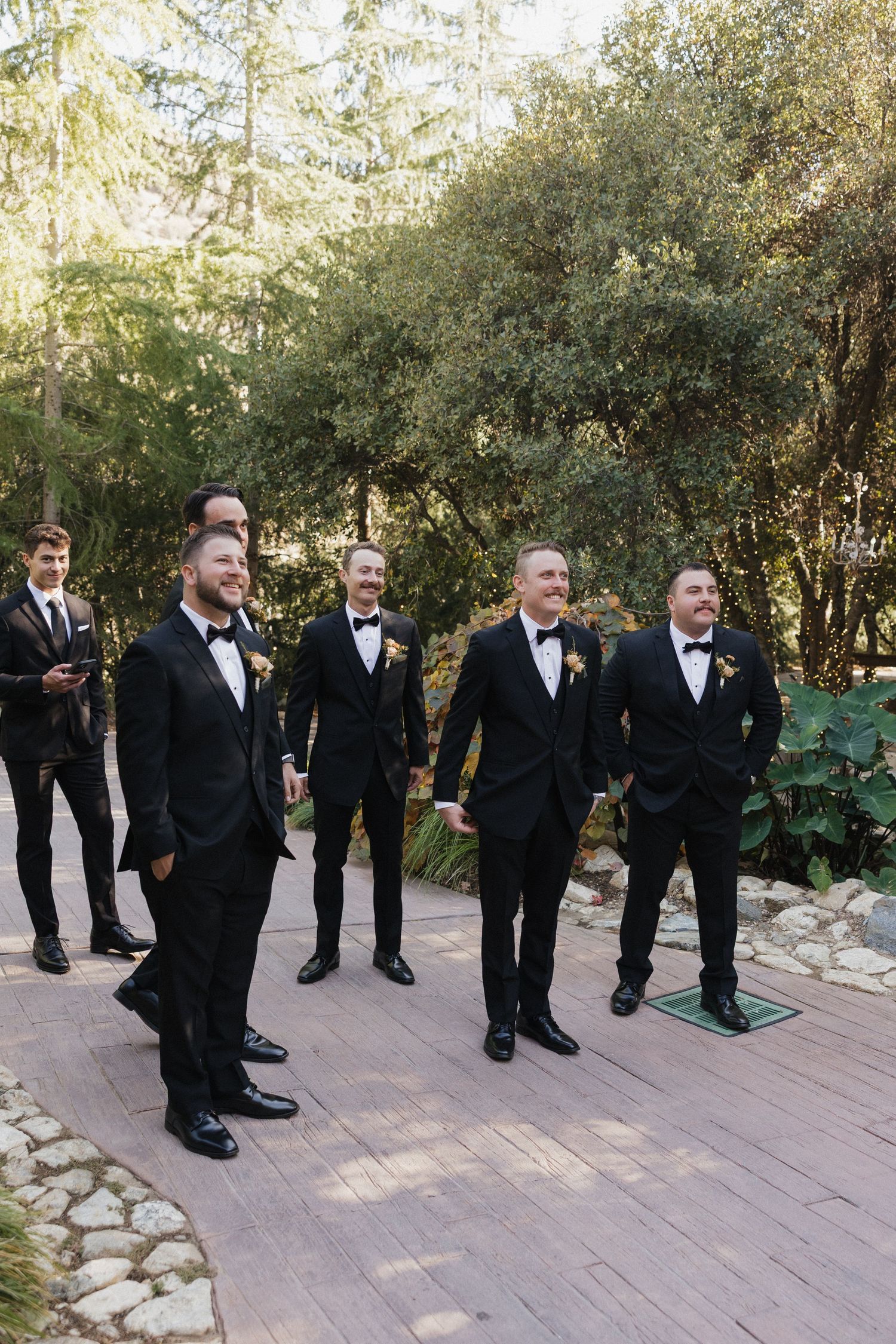 Groomsmen in black tuxedos stand in line on a stone paved pathway surrounded by greenery at an outdoor wedding venue.