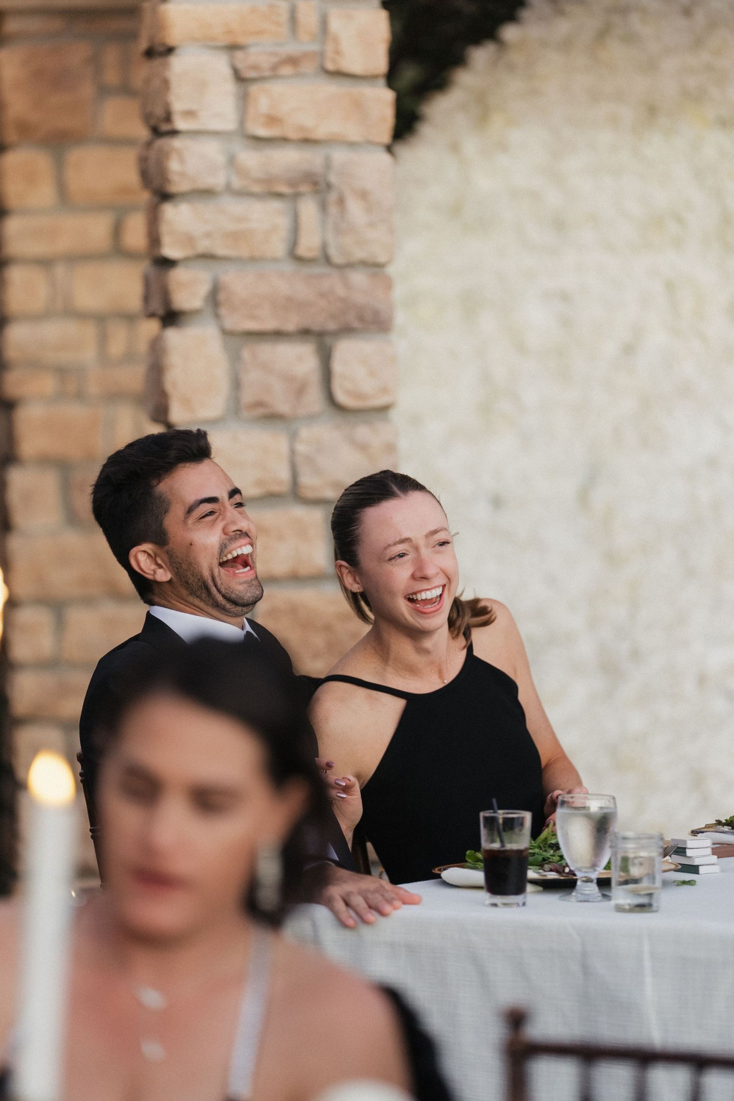 Wedding guests share genuine laughter while seated at a rustic stone venue during reception celebrations.