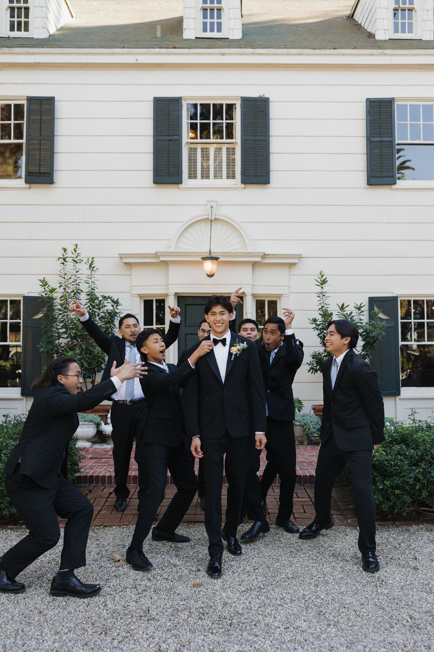 Groomsmen in black suits pose playfully in front of a white colonial building.
