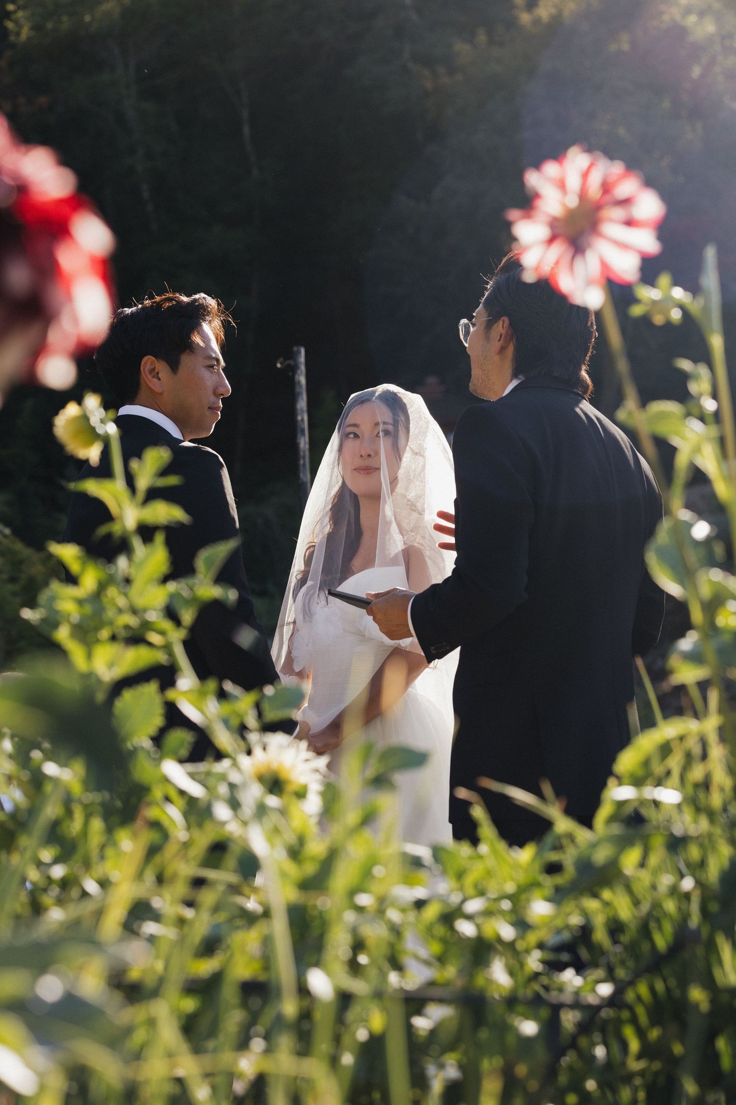 A wedding ceremony takes place in a garden with pink flowers blooming in the foreground.