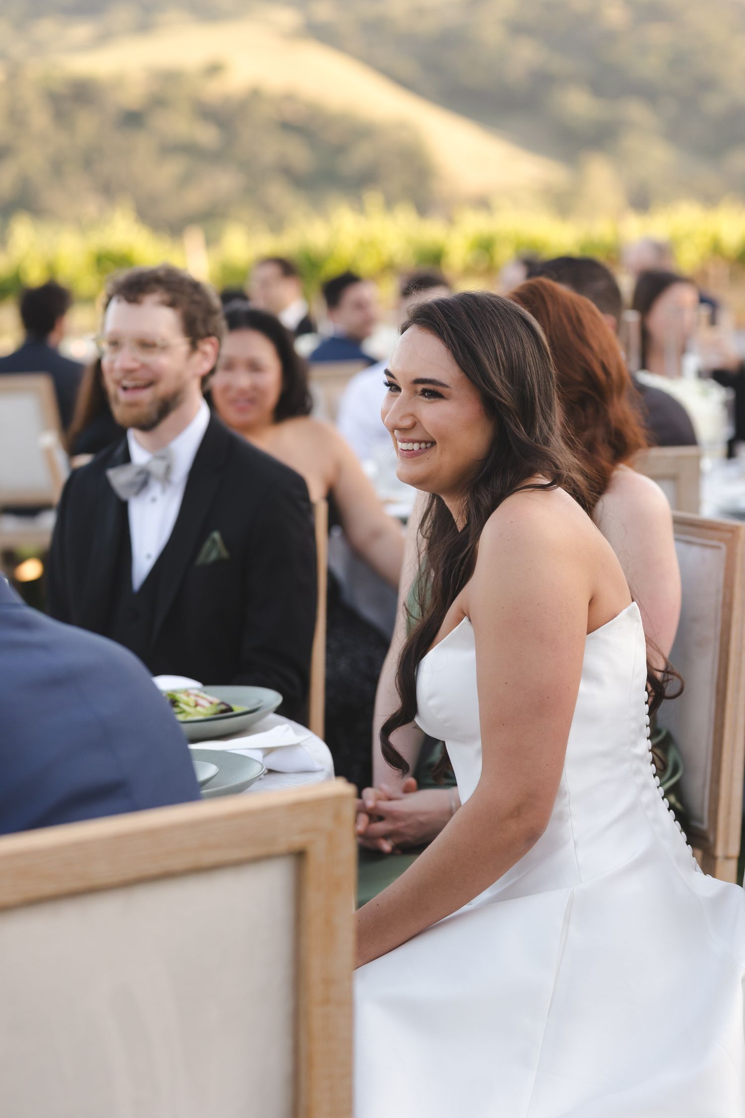 Outdoor wedding reception at sunset with guests dining among vineyard views.