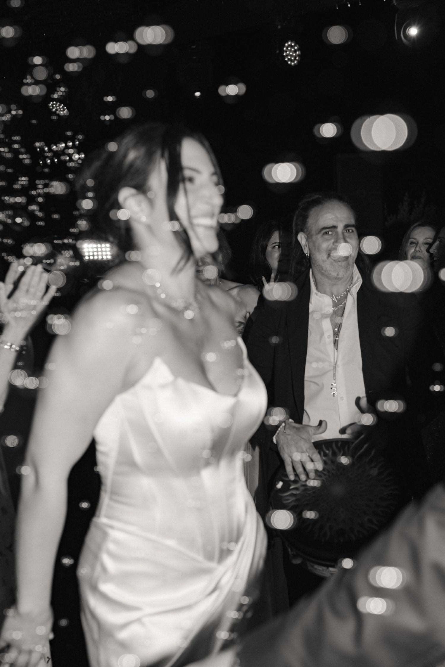 Black and white photo of bride and older man dancing joyfully at wedding reception with bokeh lights.