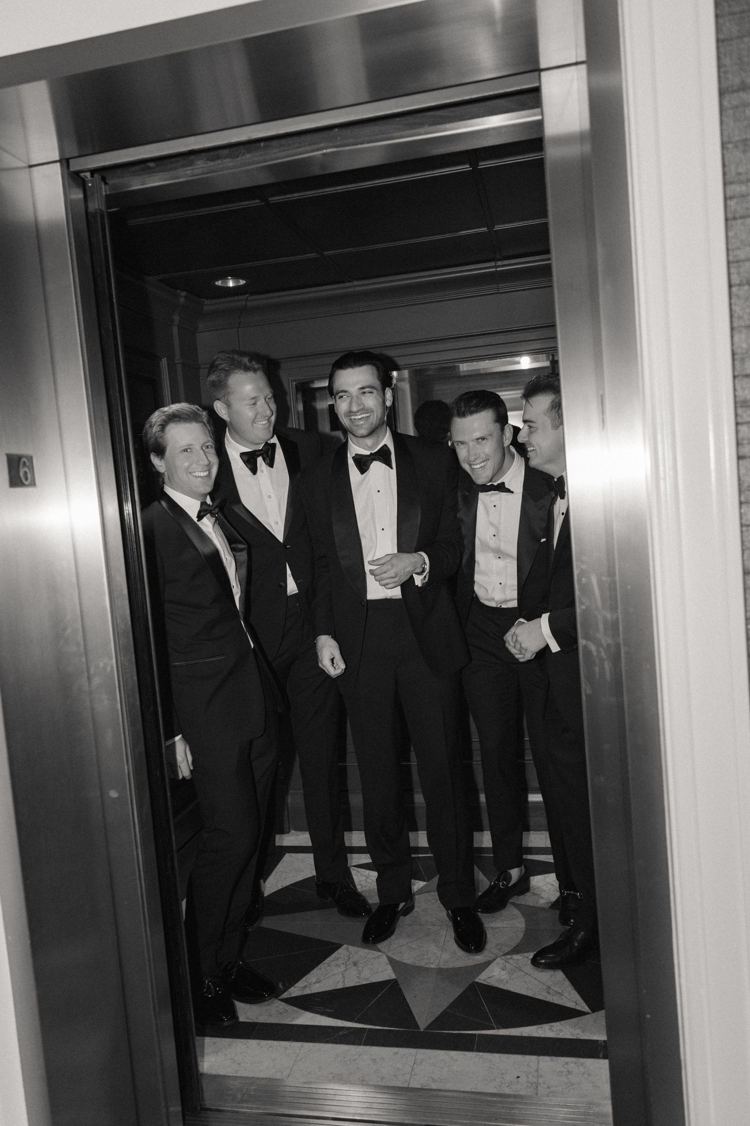 Black and white photo of four men in tuxedos smiling inside an elevator at a formal event.