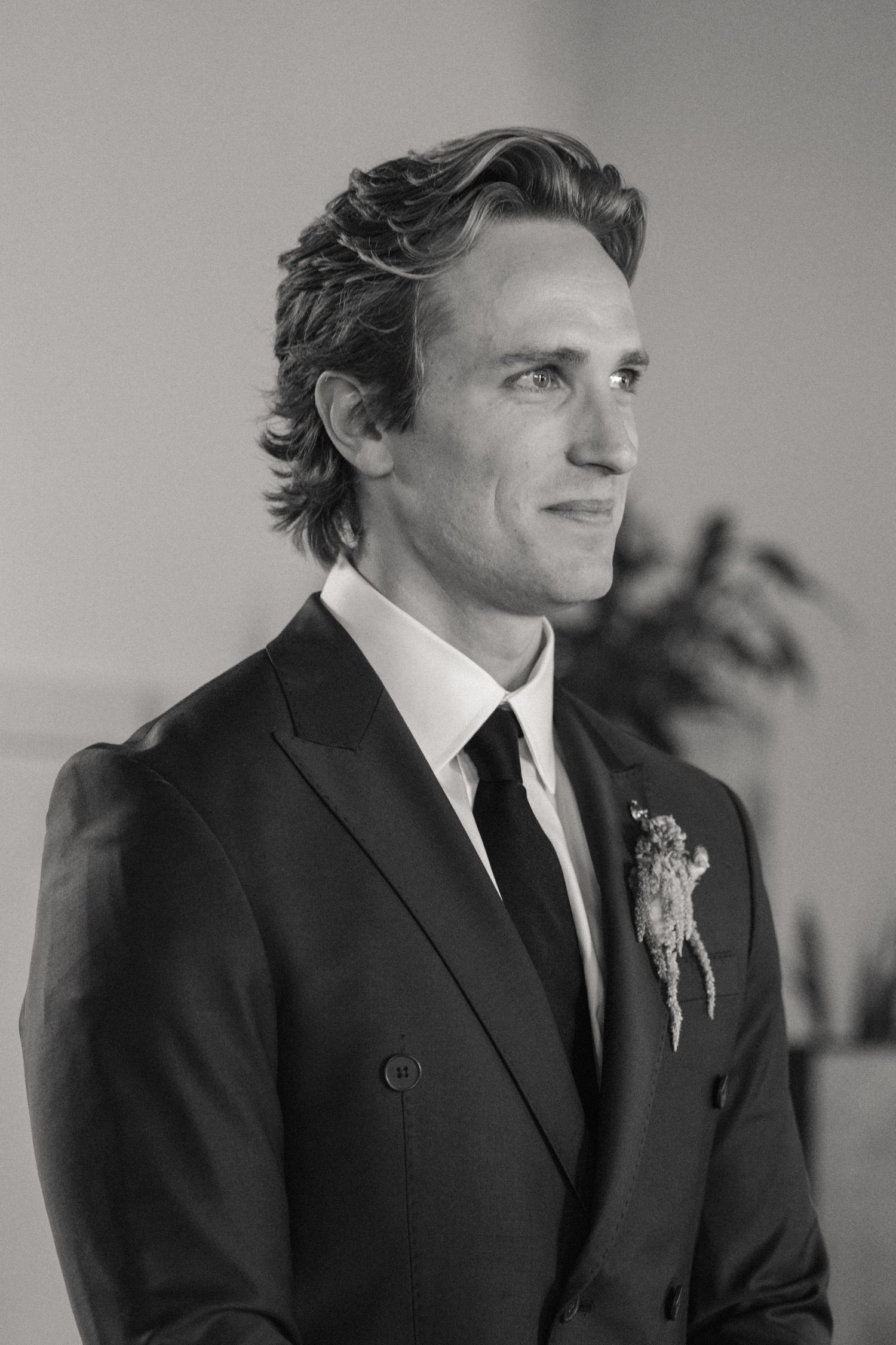 Black and white portrait of a groom in a dark suit with boutonniere, smiling softly at a wedding ceremony.