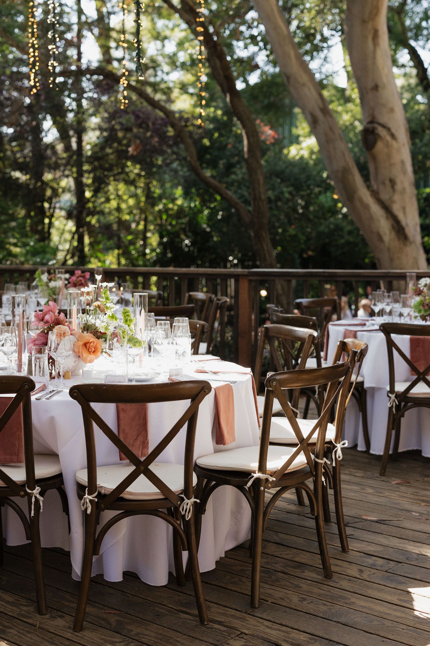 Elegant outdoor wedding reception on wooden deck with cross-back chairs, floral centerpieces, and string lights among trees.