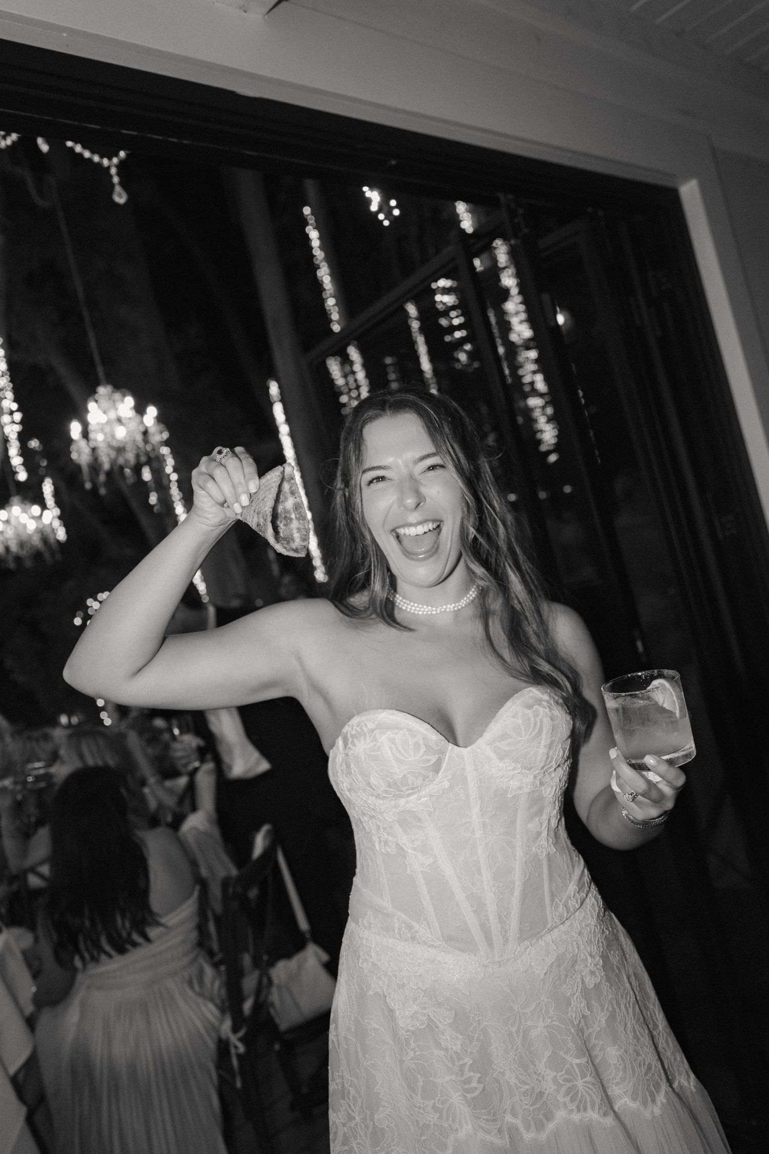 Black and white photo of an exuberant bride in lace gown dancing and holding a drink at her reception.