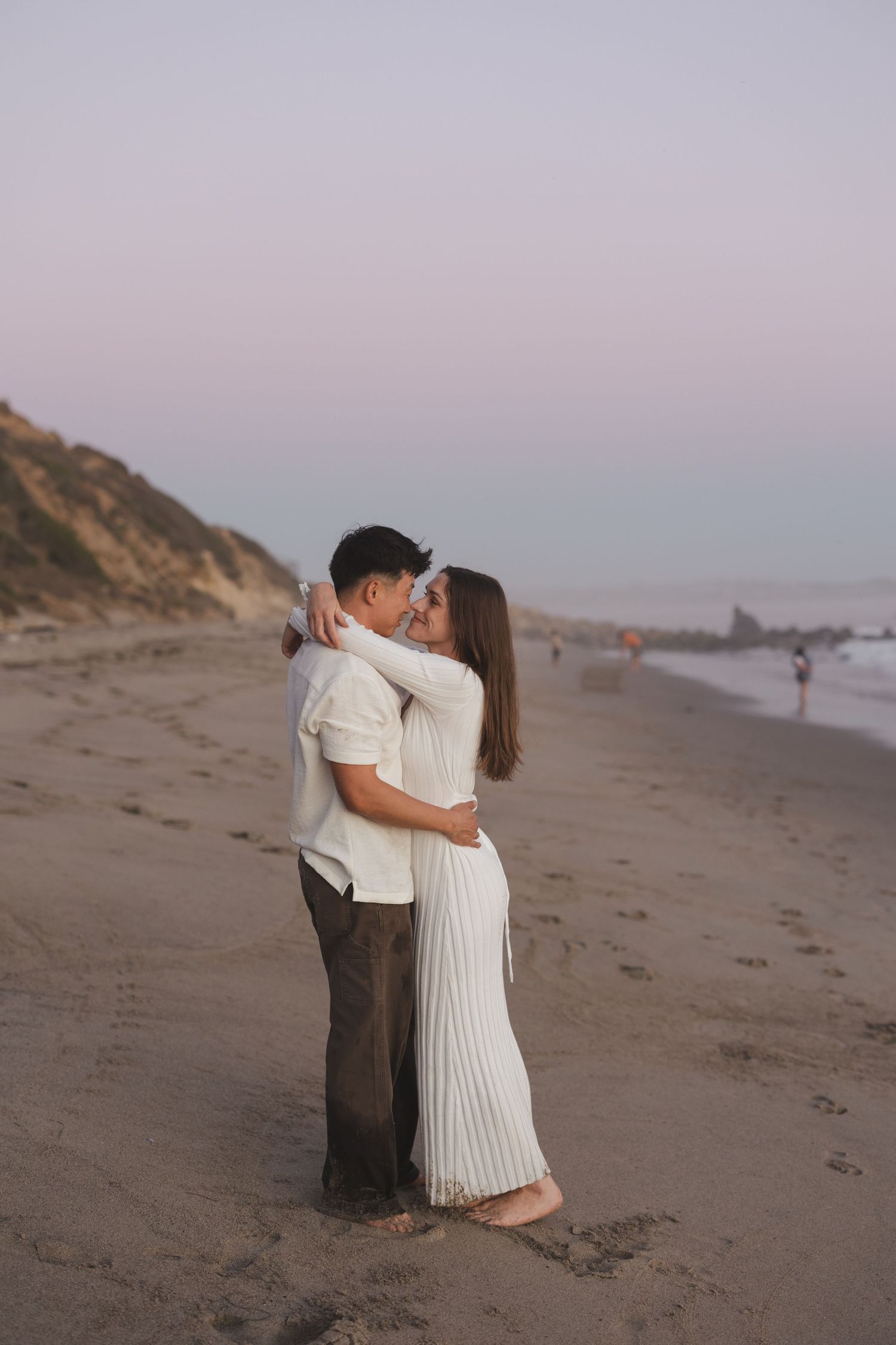 Romantic couple embraces on a misty beach at sunset with coastal cliffs and waves in the background.