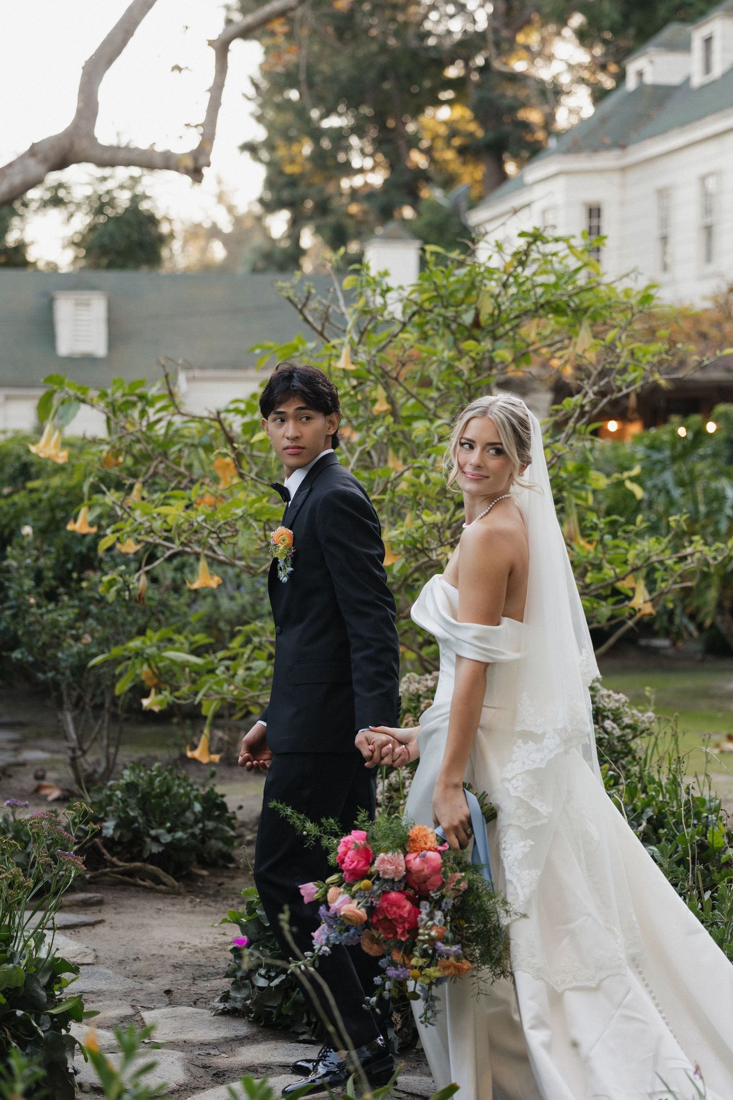 A couple poses for wedding photos in a lush garden with colorful flowers at sunset.