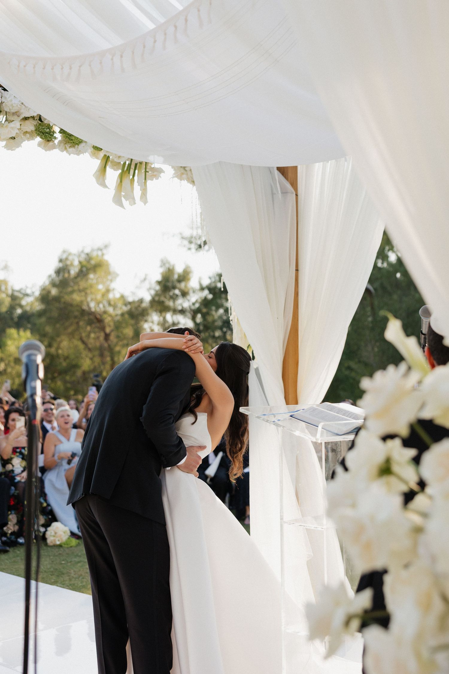 A couple shares their first kiss during an outdoor wedding ceremony under a white draped canopy with floral decorations.