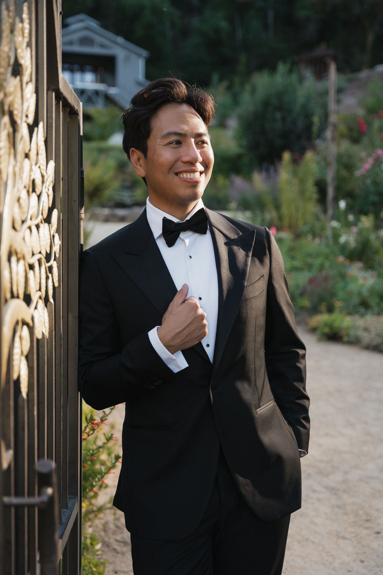 A groom in a classic black tuxedo poses against a stone wall in a garden setting.