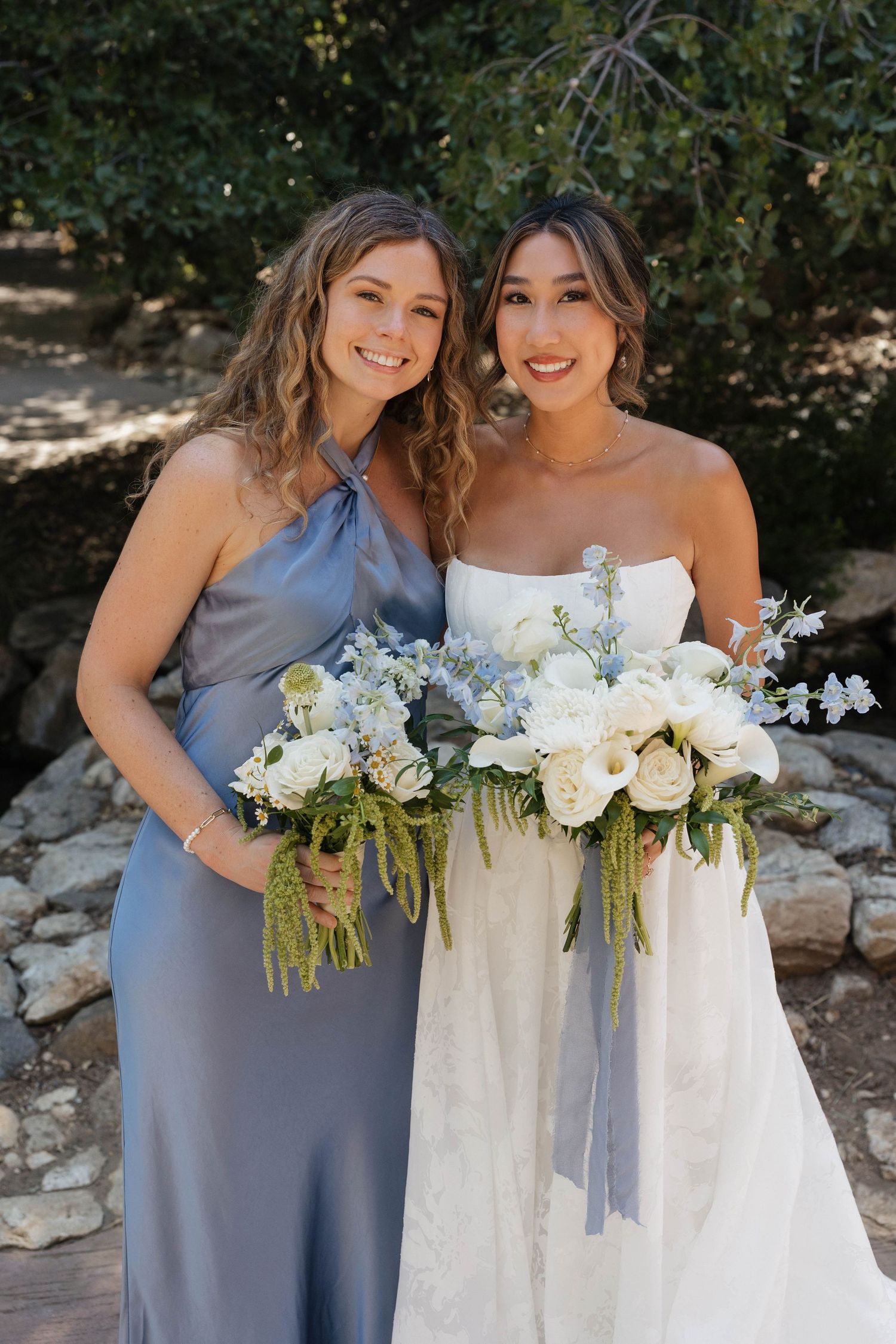 Bridesmaid in blue and bride holding elegant white and blue floral bouquets outdoors.