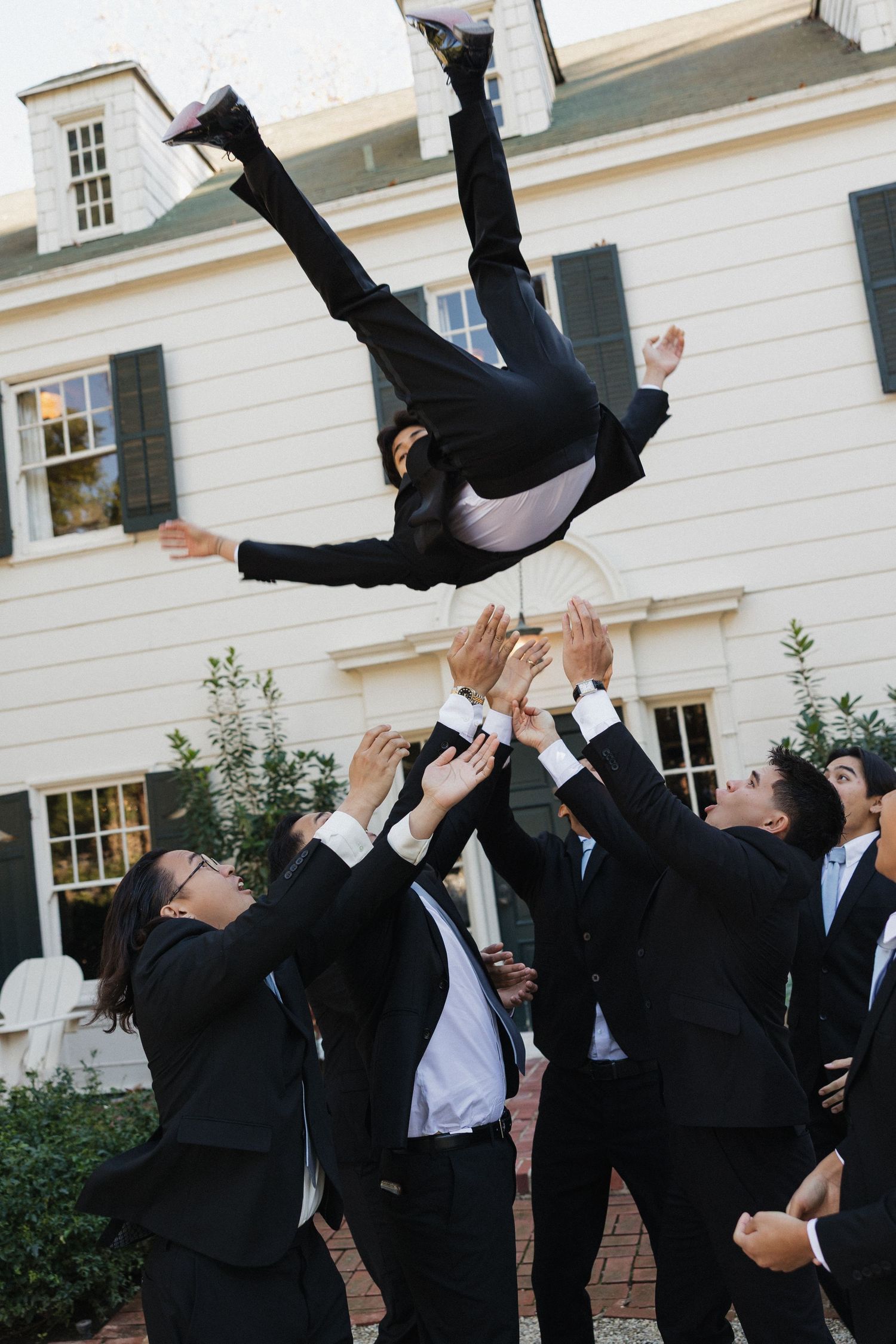 Groomsmen toss another groomsman in the air in a celebratory moment outside a white building.