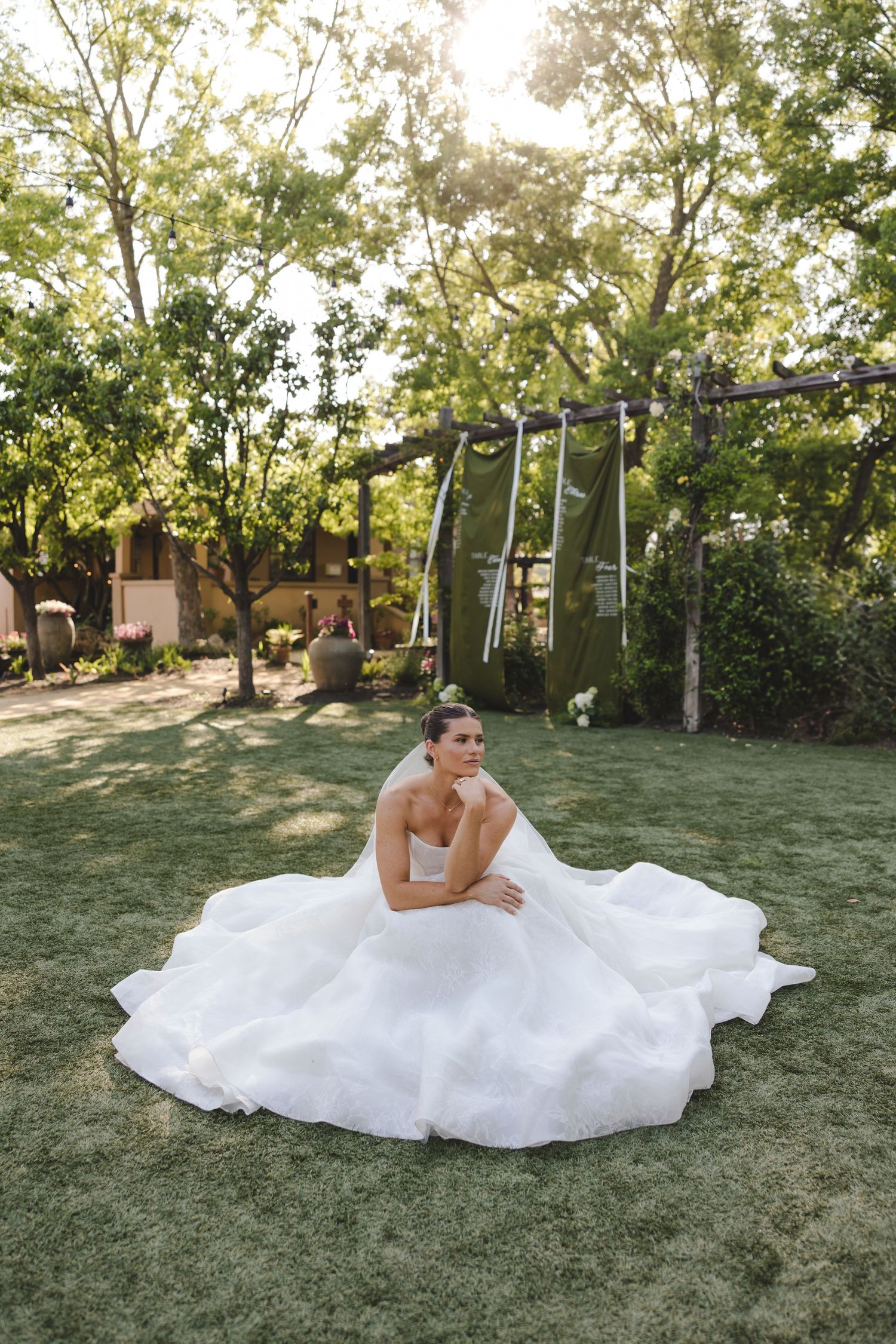 A bride in a white ball gown sits gracefully on a lawn with trees and a swing set in the background at sunset.