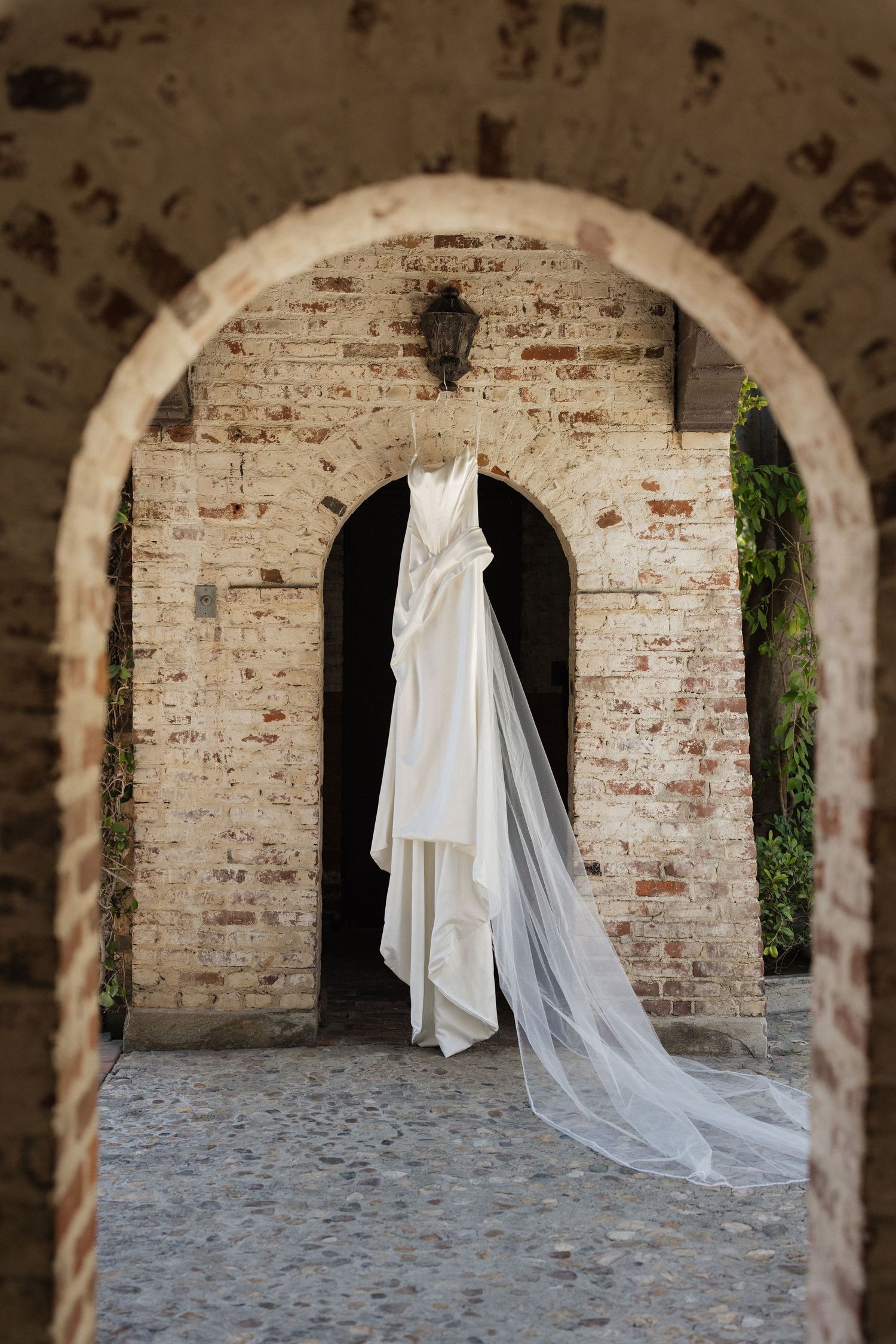 Elegant white wedding dress with long veil hanging in a rustic brick archway doorway of a historic stone building.
