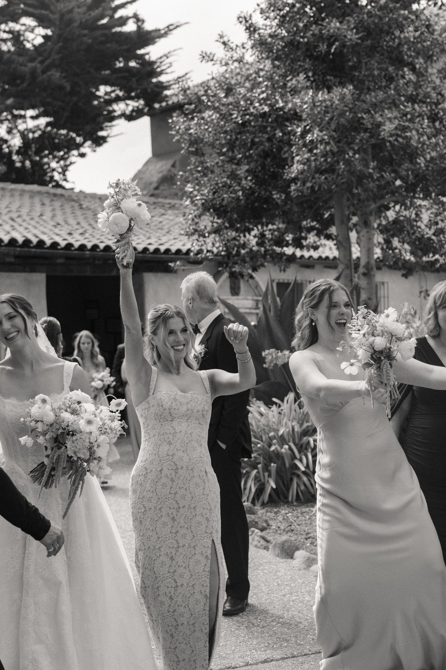 Black and white photo of joyful bride celebrating with bouquet raised, surrounded by bridesmaids at outdoor wedding ceremony.