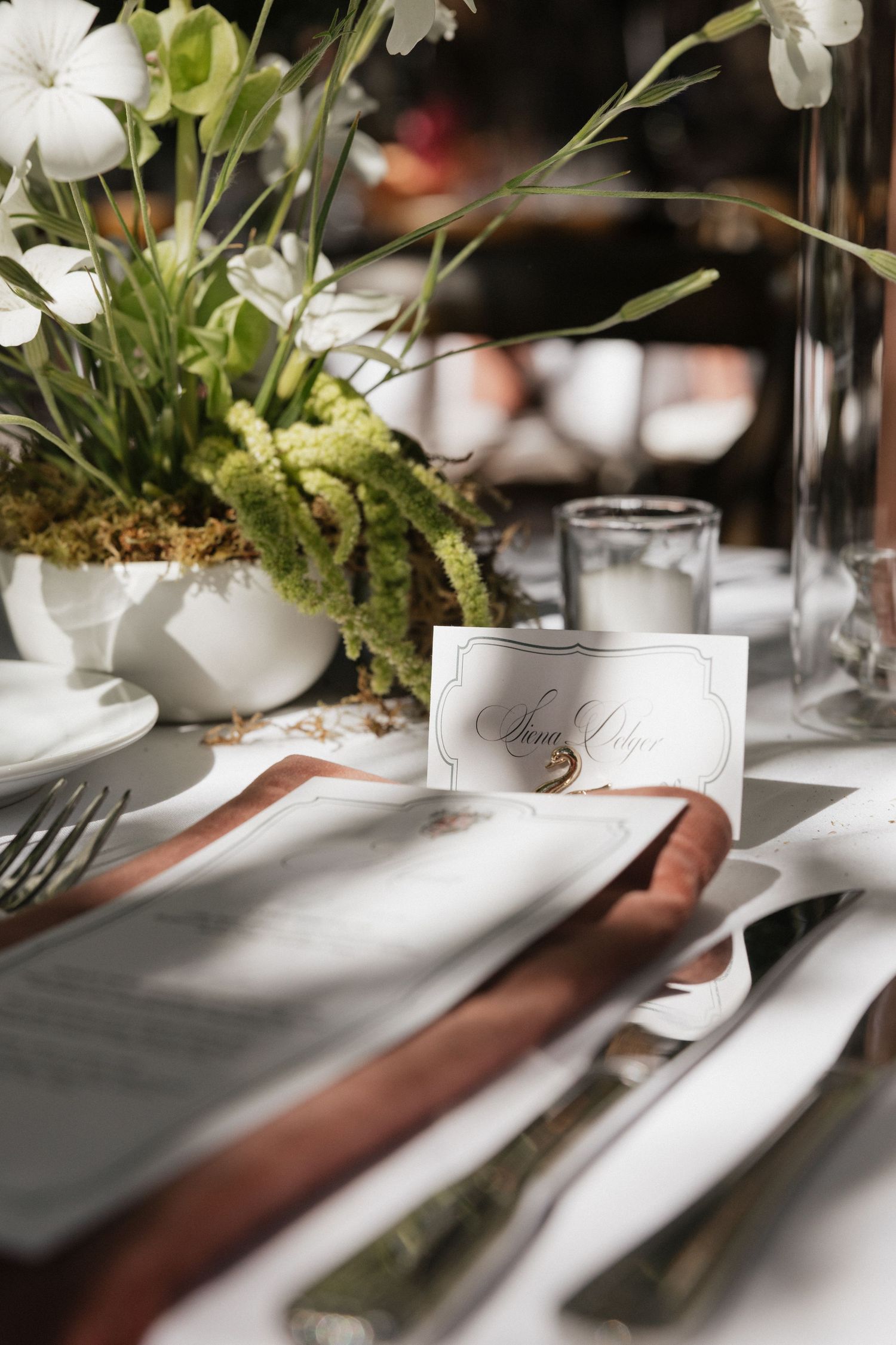 Elegant wedding place setting with calligraphy name card, copper napkin ring, silver cutlery, and white floral centerpiece.