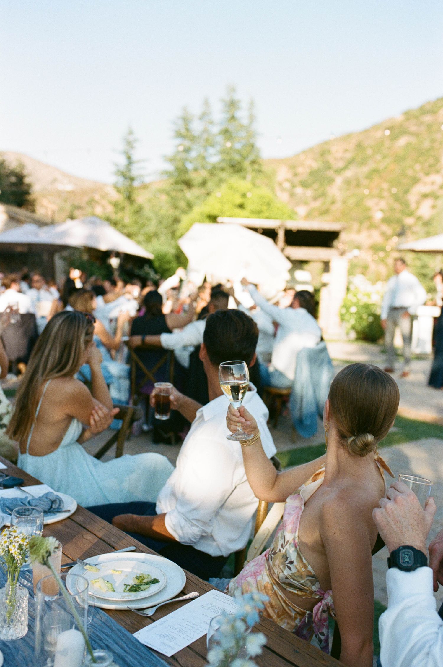 Outdoor wedding reception with guests dining at decorated tables in a mountain setting.