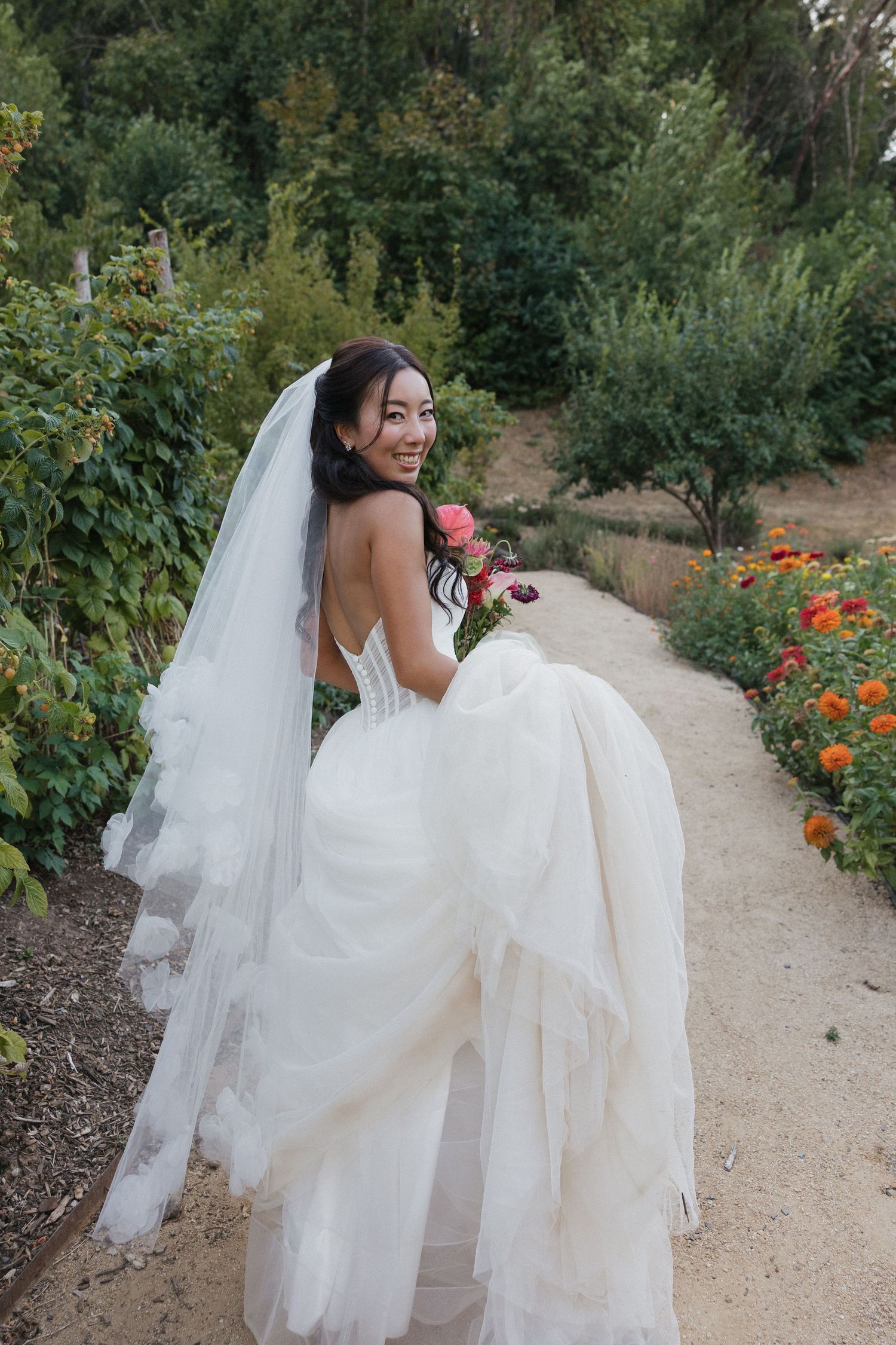 A bride in a flowing white dress twirls joyfully in a garden path with orange flowers.