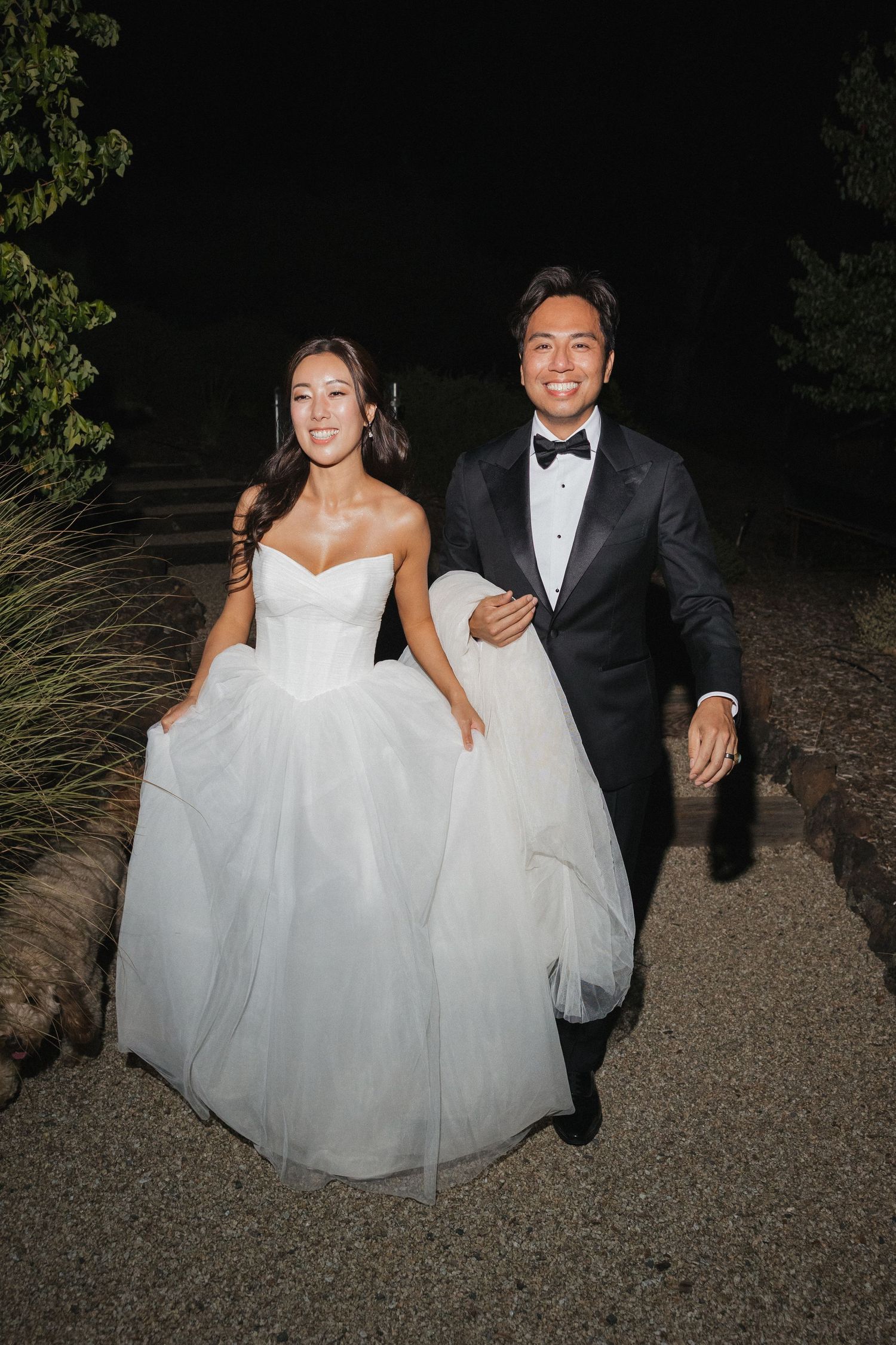 A newlywed couple walks together at night, both dressed in formal wedding attire.