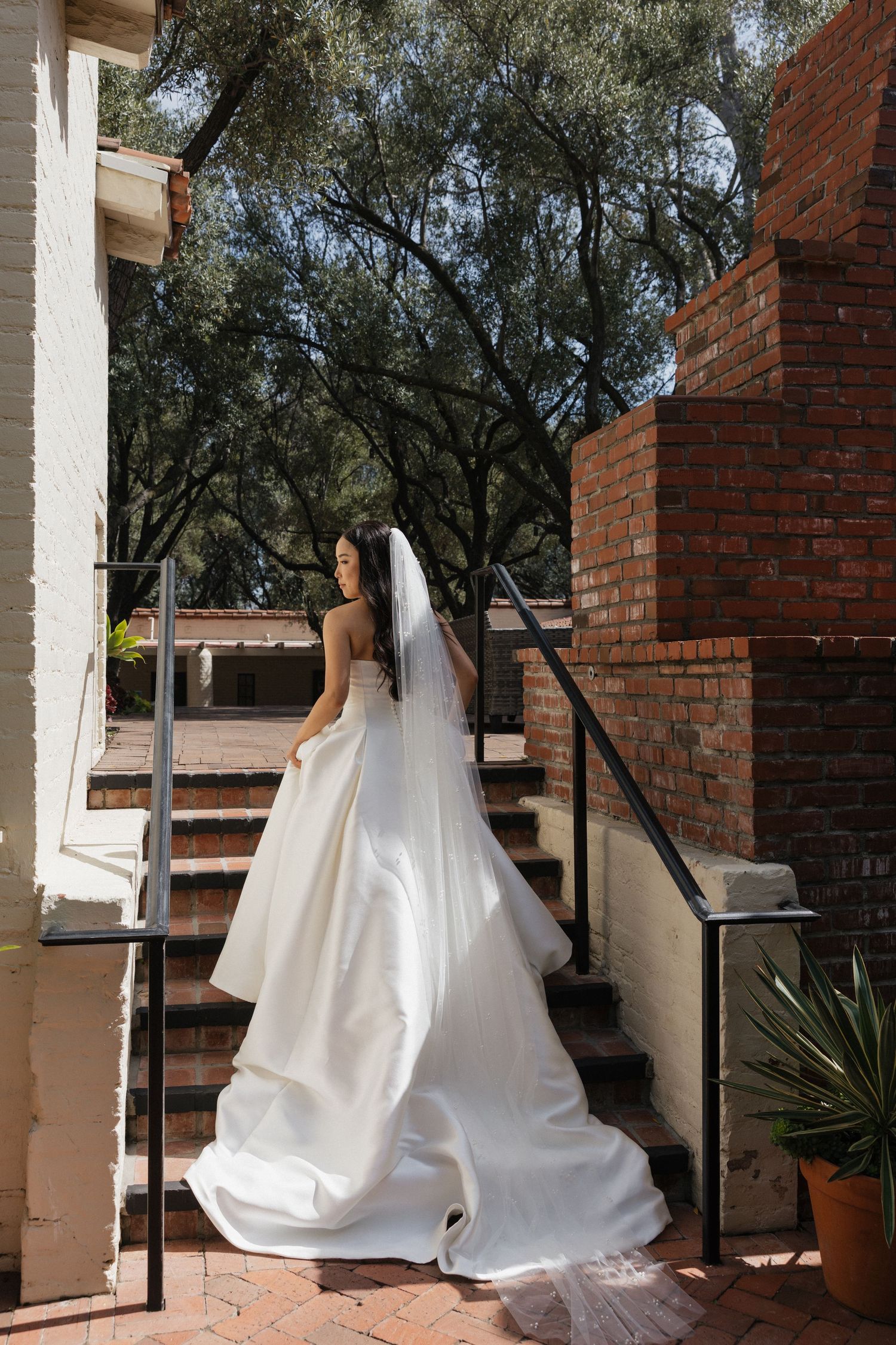 Bride in elegant white ball gown with veil stands on brick staircase surrounded by lush green trees.