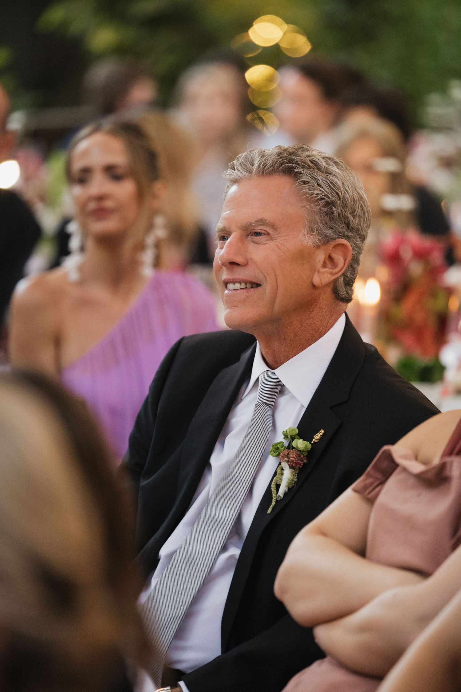 Smiling older man in black suit with boutonniere seated at an elegant outdoor wedding reception.