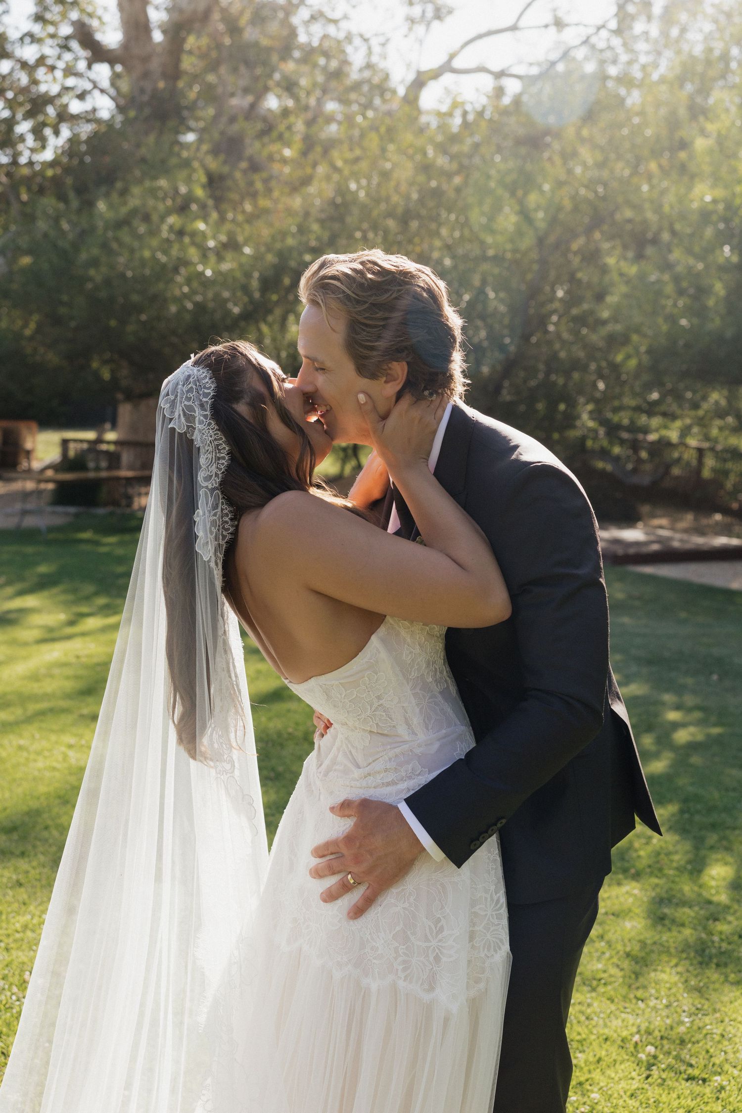 Bride and groom sharing first kiss outdoors in sunny garden on wedding day.