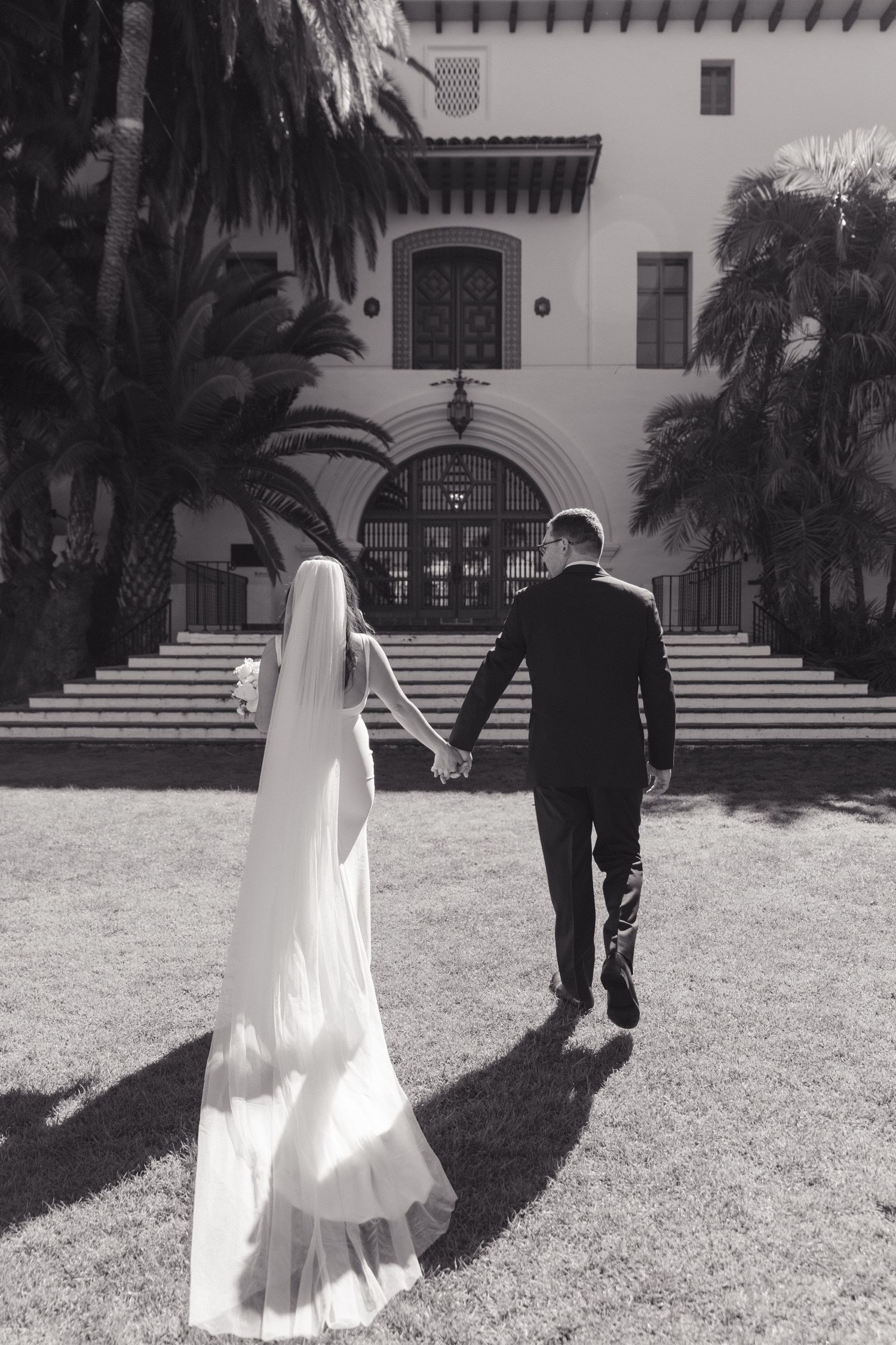 Black and white wedding photo of newlyweds holding hands and walking towards ornate Spanish-style building entrance.