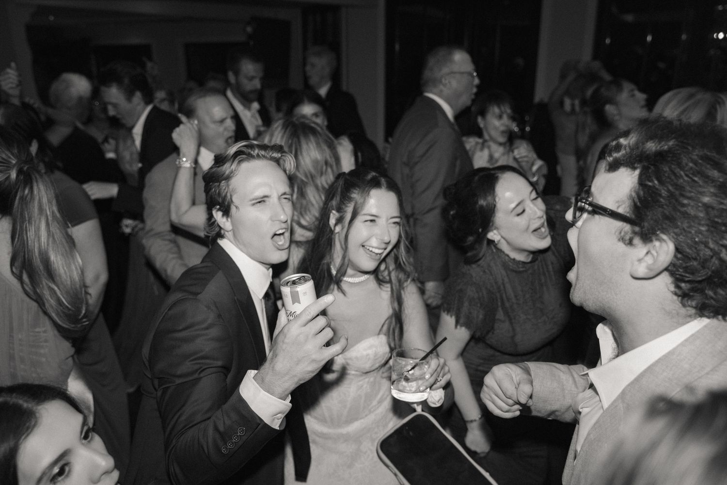 Black and white photo of bride and groom laughing with guests on lively wedding dance floor.