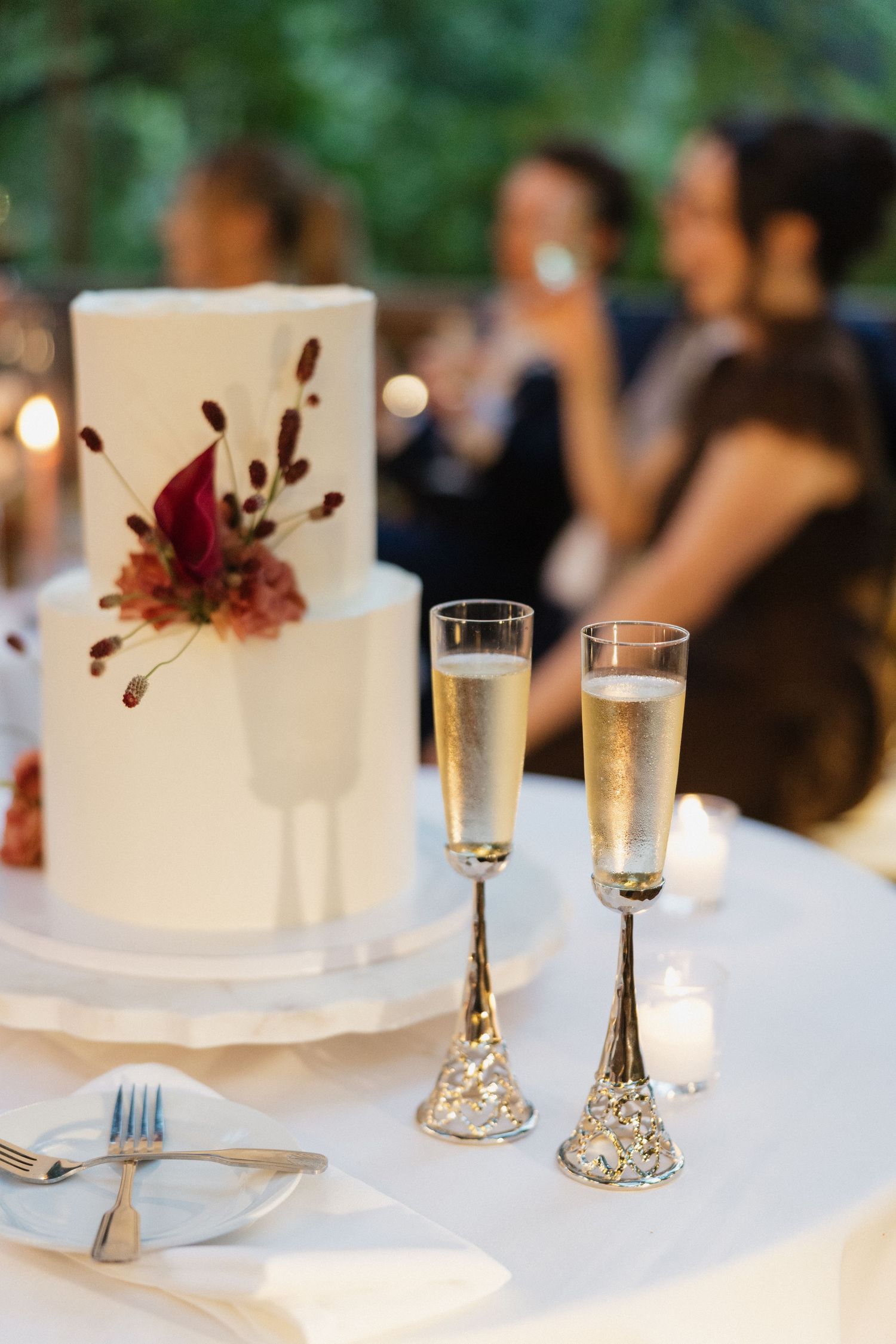 Two ornate gold champagne flutes beside a white two-tier wedding cake decorated with burgundy and pink florals.
