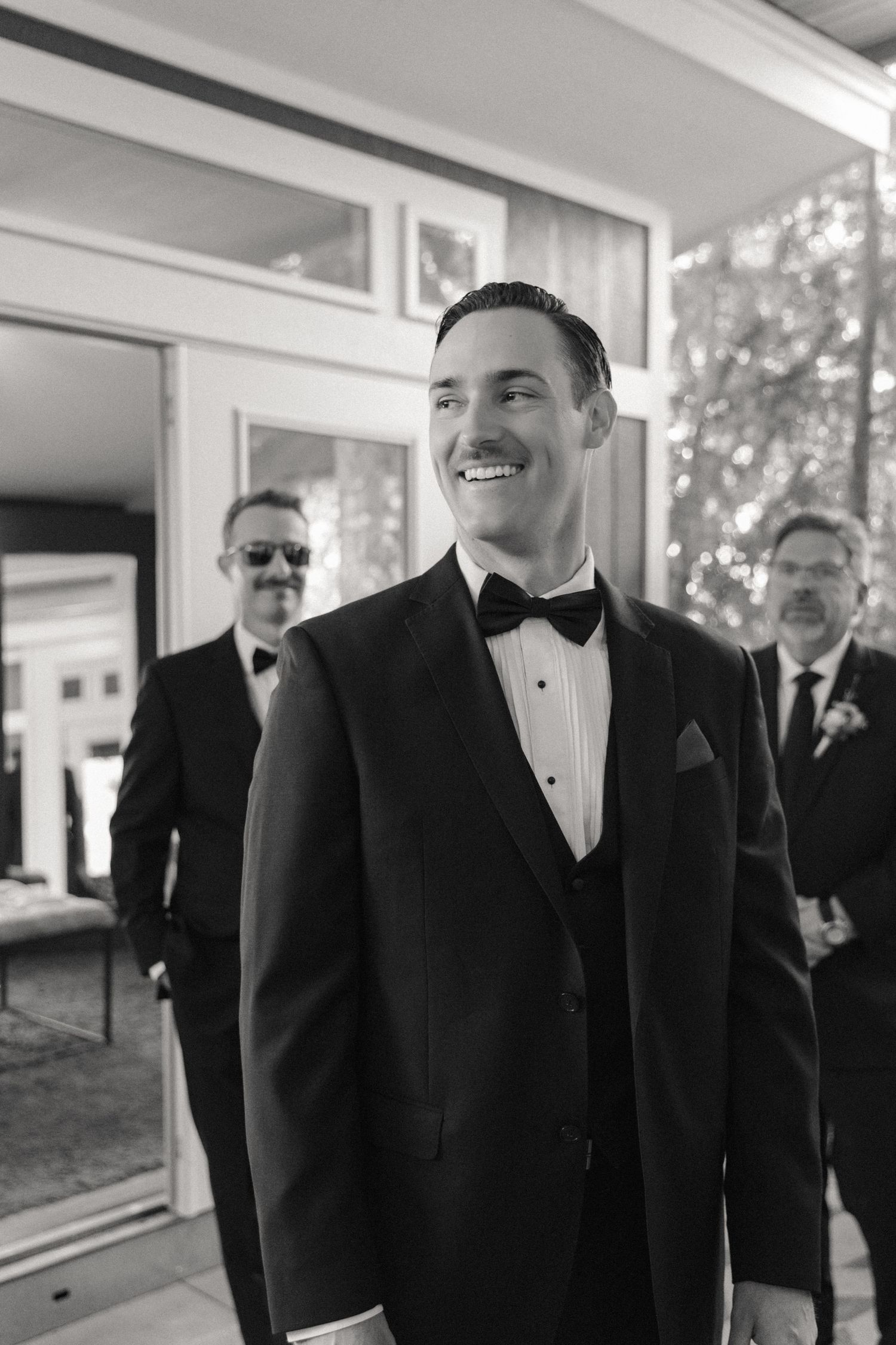 Group of formally dressed people in black tuxedos and bow ties standing in a bright indoor space.