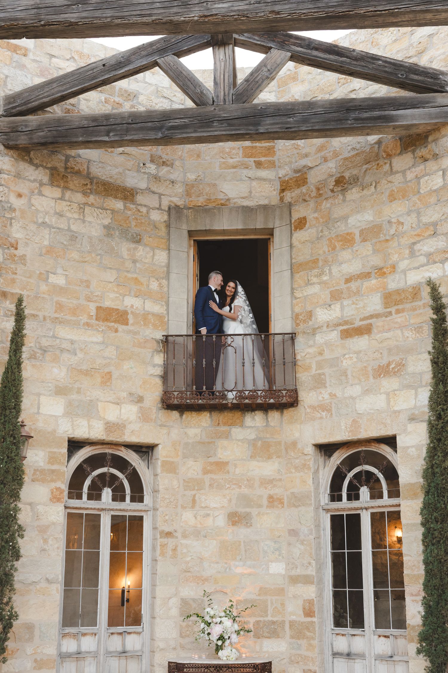 Couple stands on balcony of stone building with wooden beams and arched windows.