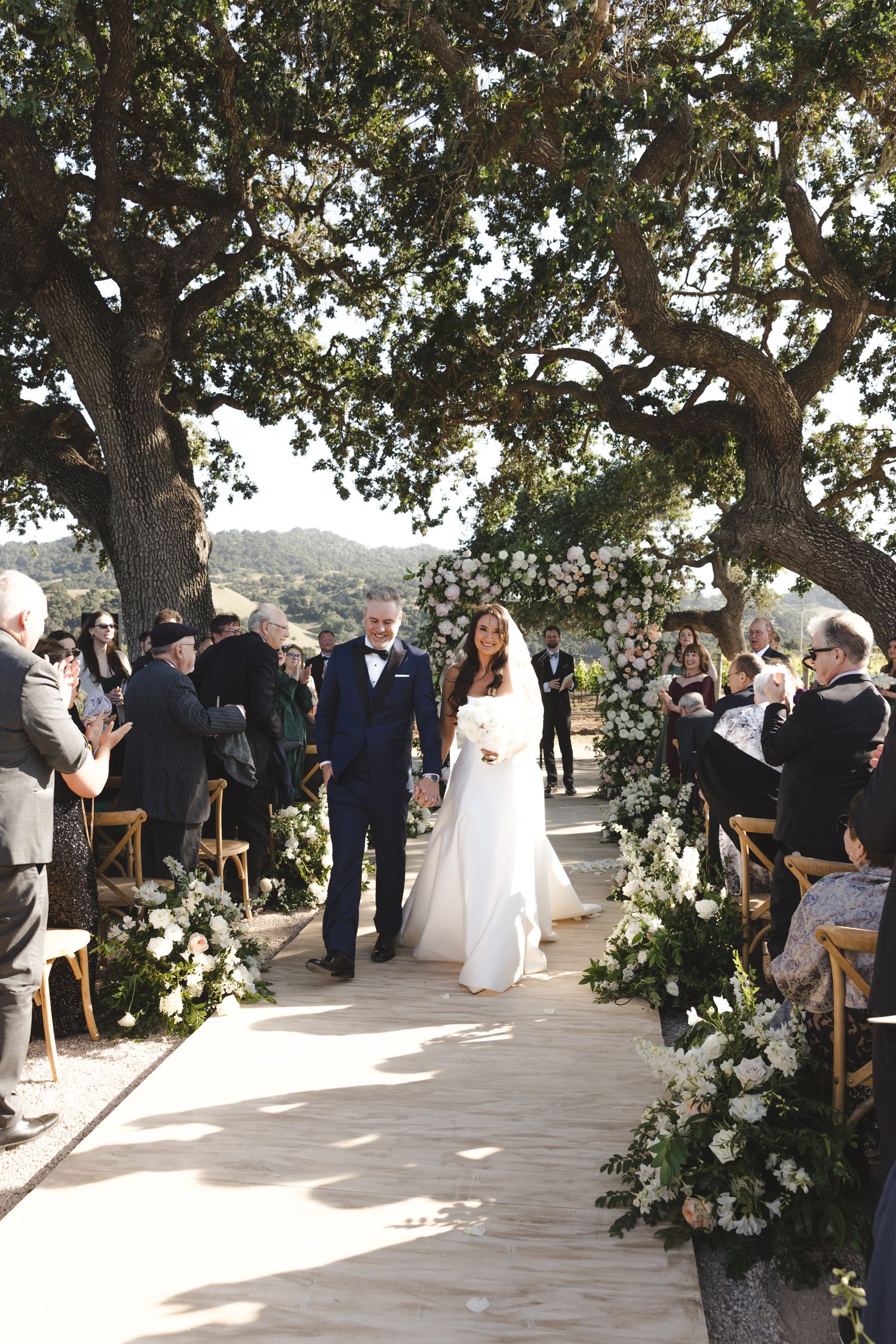 Outdoor wedding ceremony setup with wooden chairs and floral decorations under majestic trees.