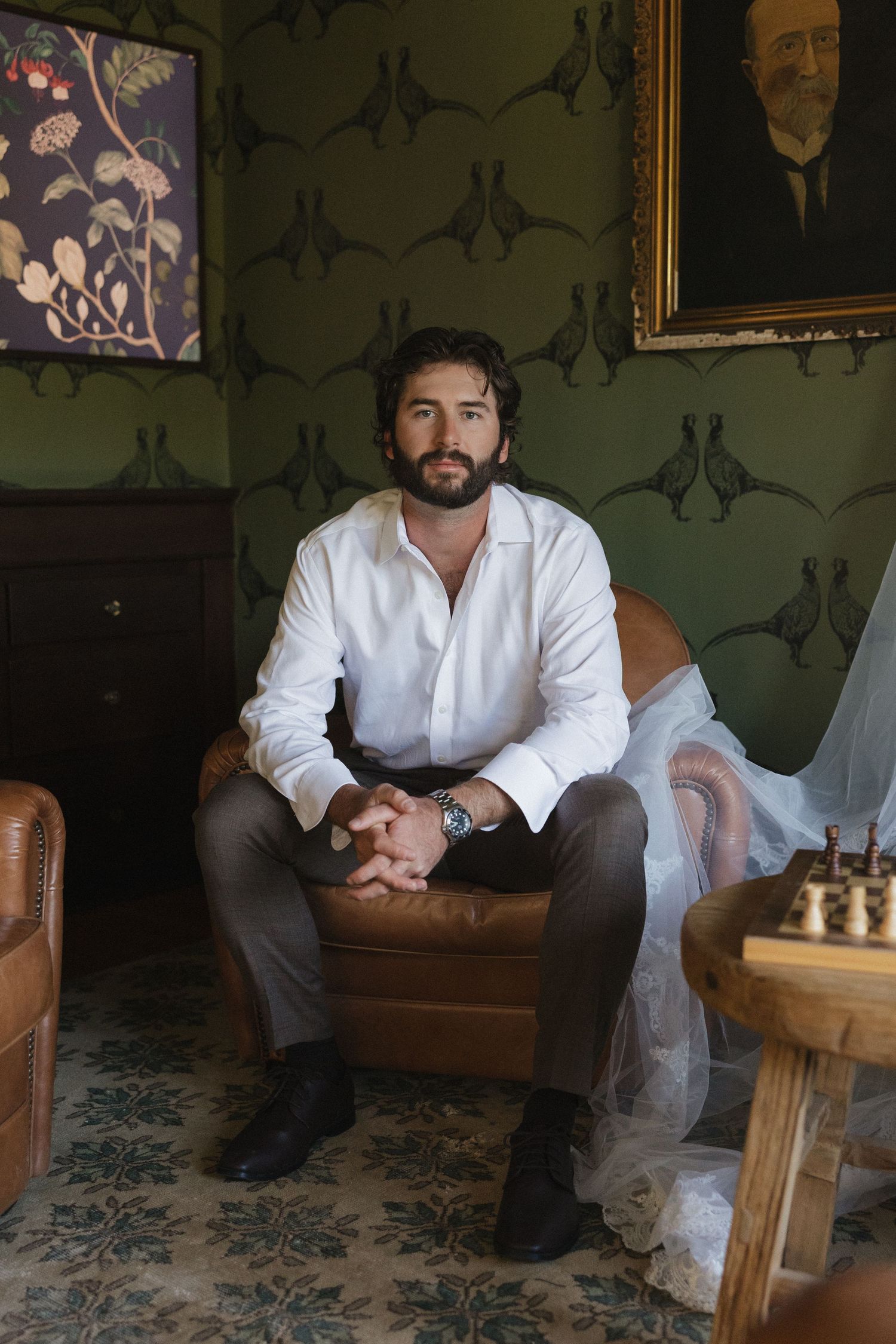A person in a white shirt sits on a brown leather couch in a room with patterned wallpaper and framed artwork.